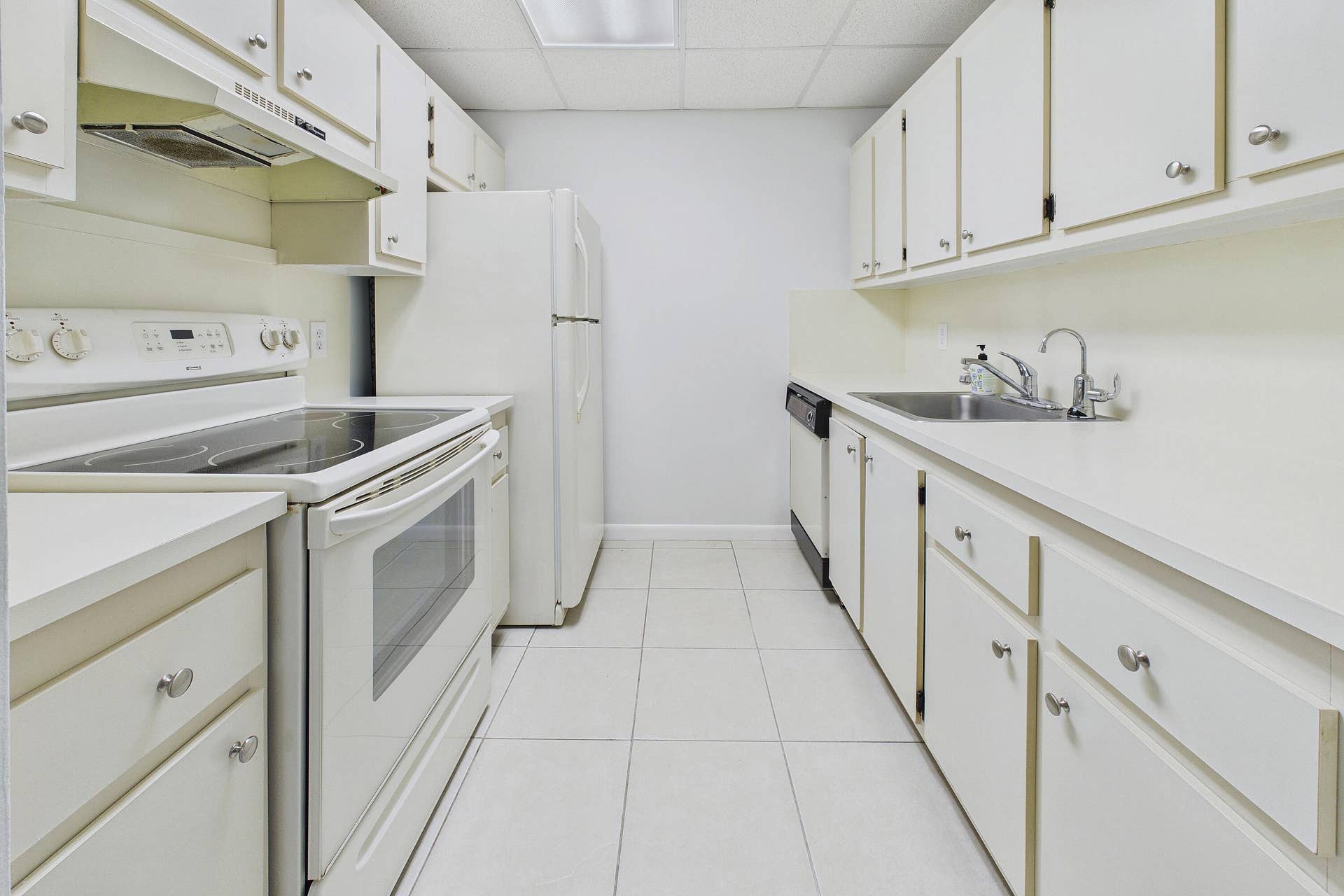13155 SW 7TH Court, #103E, Pembroke Pines, FL 33027 Galley kitchen featuring white cabinetry, solid surface countertops, tile flooring, a stainless steel sink, and white appliances