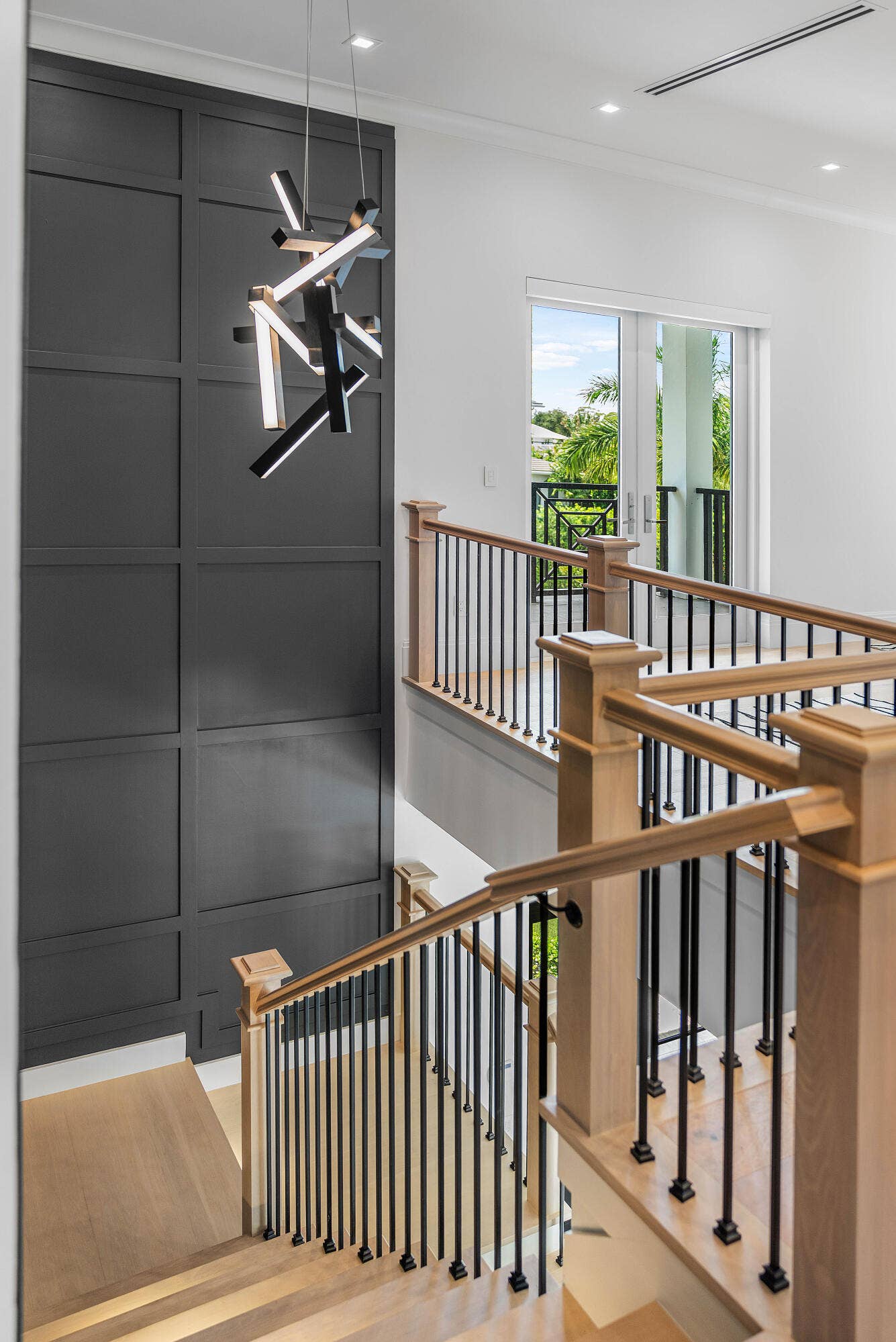 12898 S Shore Drive, Palm Beach Gardens, FL 33410 Modern chandelier illuminating a paneled accent wall, light wood-finish stairs, and black metal balusters with wood handrails