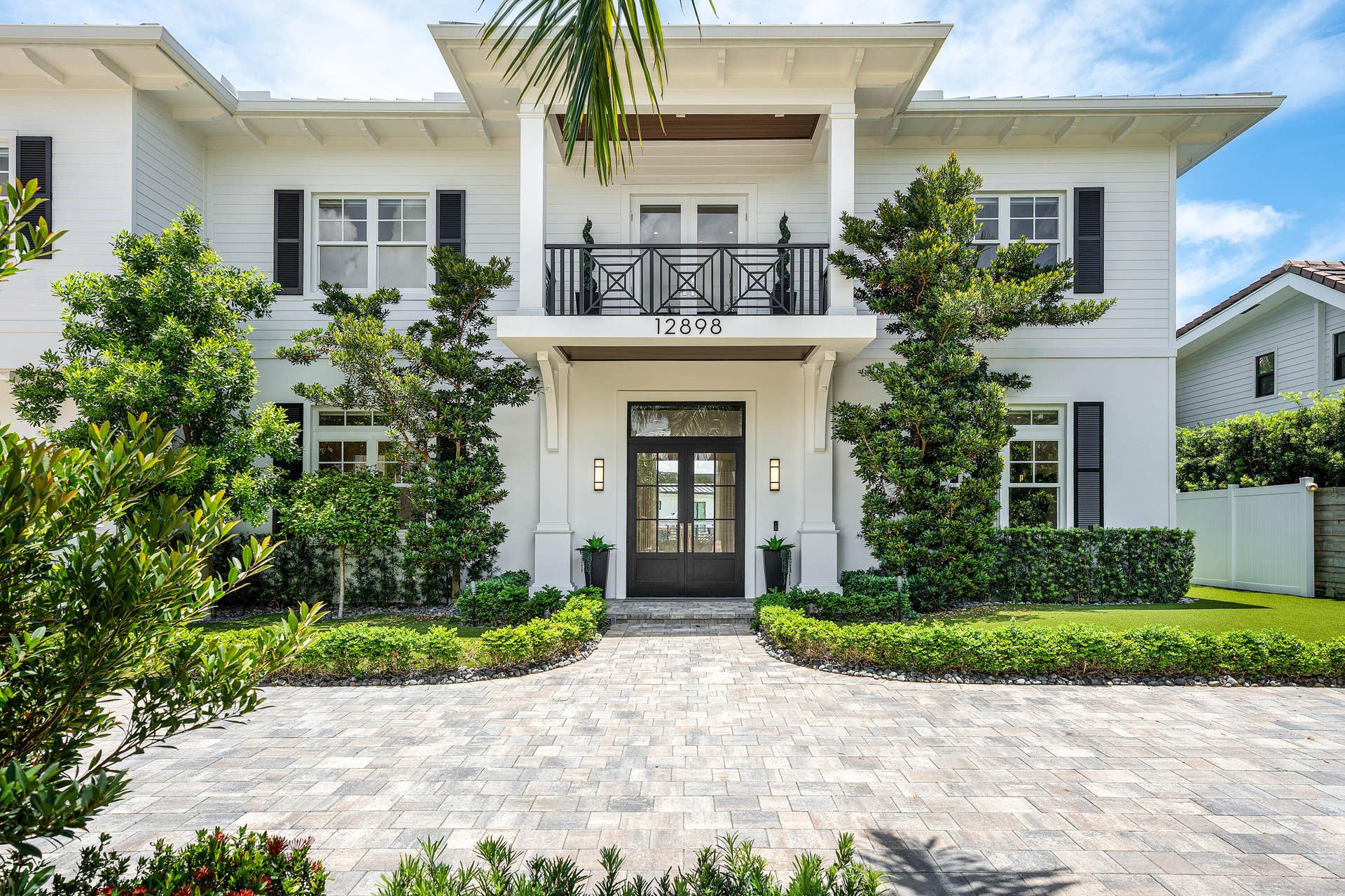 12898 S Shore Drive, Palm Beach Gardens, FL 33410 White exterior with black shutters, double-door entry, a second-story balcony with an iron railing with mature landscaping.