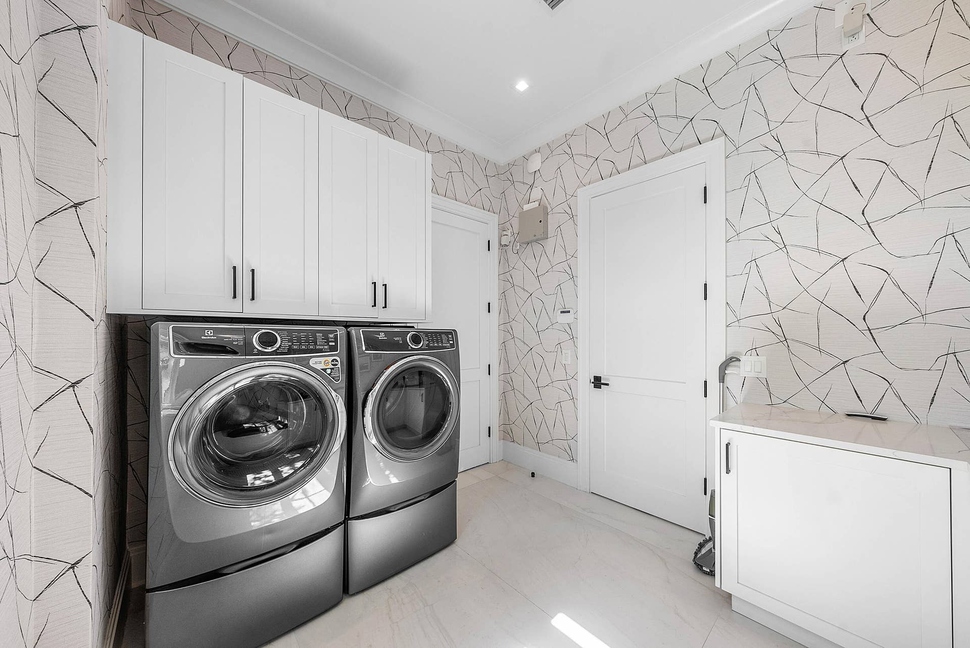 12898 S Shore Drive, Palm Beach Gardens, FL 33410 Ground Level Laundry Room with upper cabinetry with matte black hardware, white panel doors, graphic pattern wallpaper, and a built-in lower cabinet with a light-toned countertop
