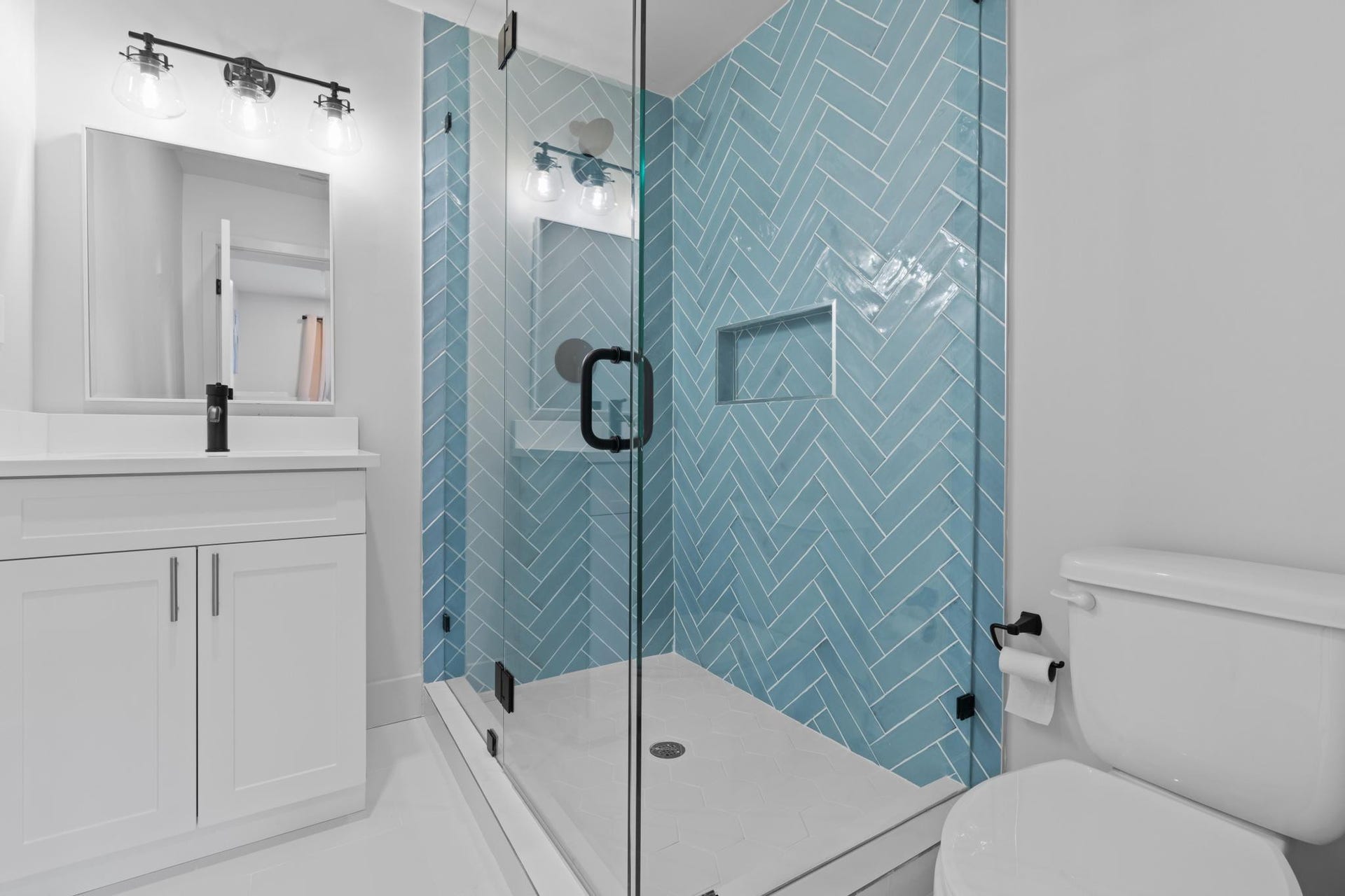 Glass-enclosed shower with herringbone tile, white vanity with integrated sink, and modern black fixtures