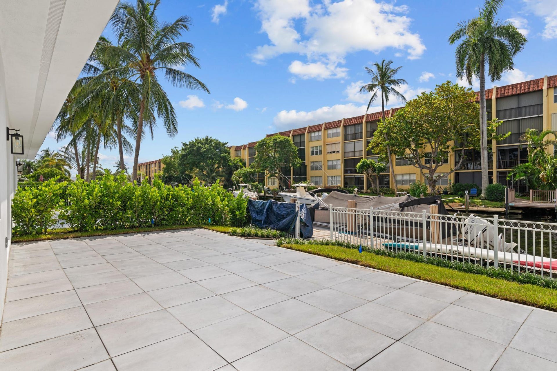 Expansive patio with large format pavers, bordered by lush landscaping and a white perimeter fence, overlooking a canal with boat docks