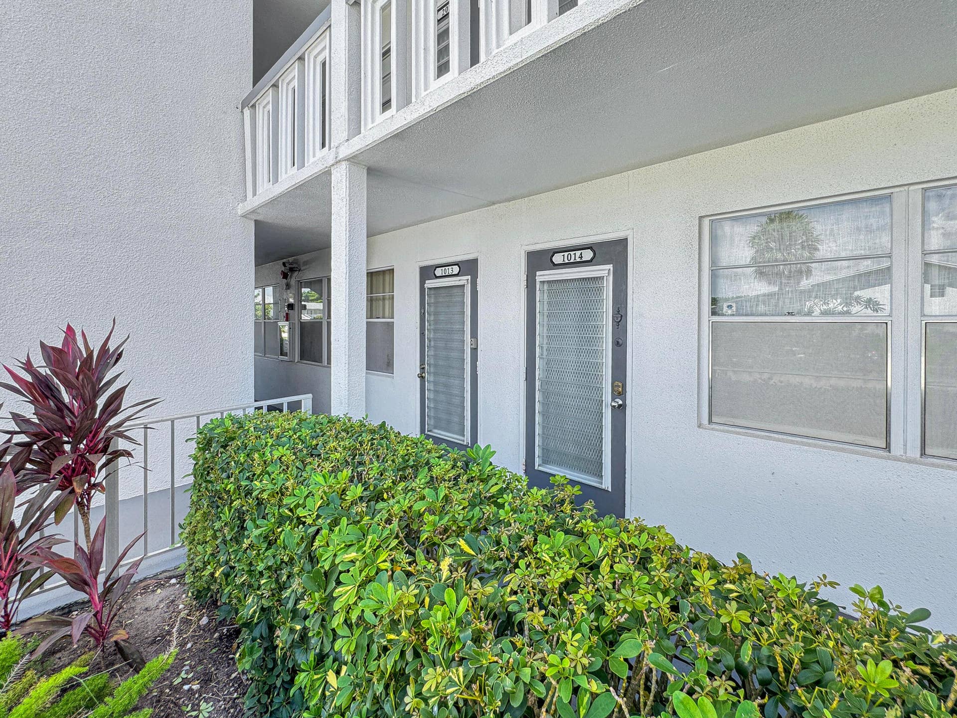 1013 HARWOOD C, #1013, Deerfield Beach, FL 33442 Building exterior with a textured finish, balcony with white railings, and manicured landscaping featuring various shrubs and plants