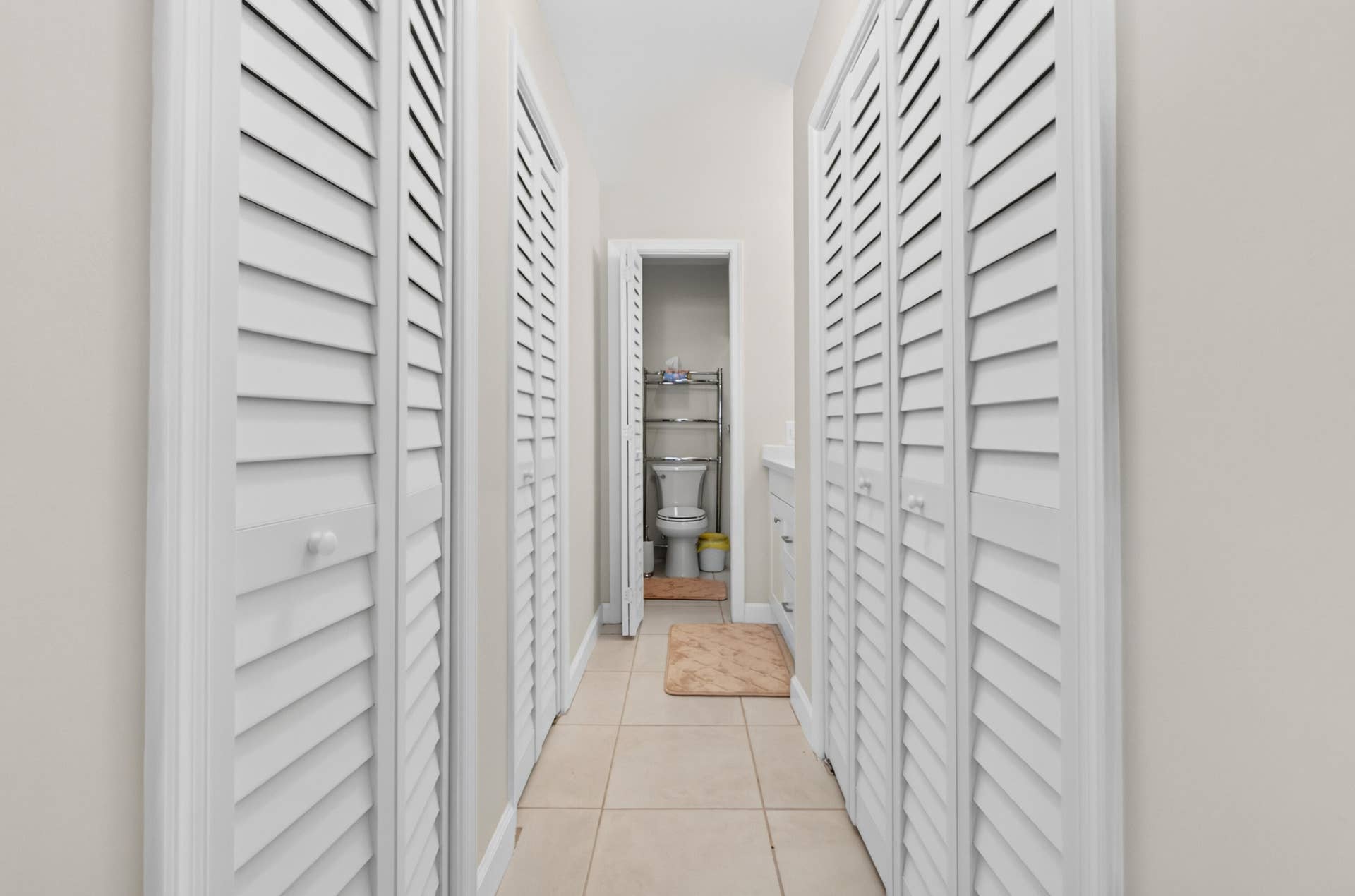 6359 Bay Club Drive, #2, Fort Lauderdale, FL 33308 Hallway featuring white louvered closet doors on either side, neutral tile flooring, and bright overhead lighting creating a clean, organized passage to the adjacent bathroom