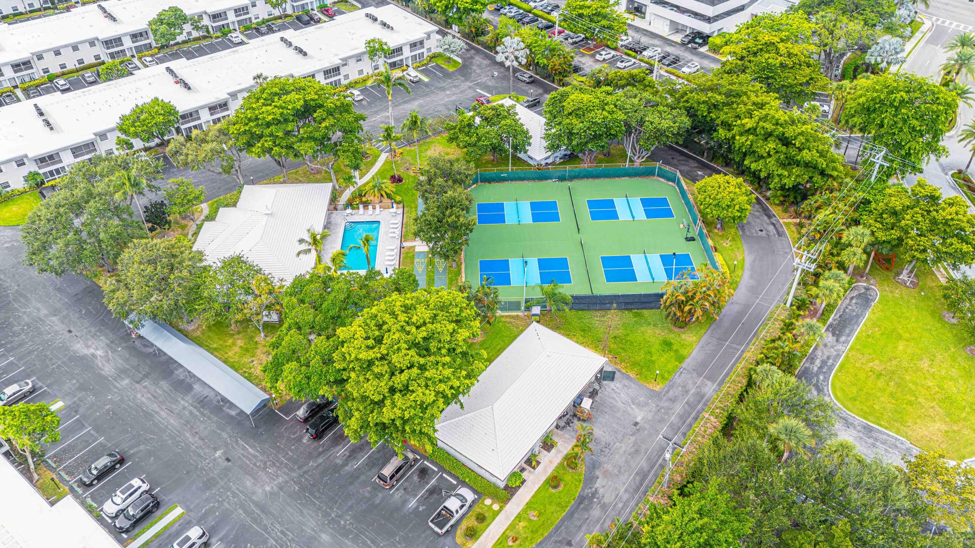 6359 Bay Club Drive, #2, Fort Lauderdale, FL 33308 Aerial view of a recreational facility featuring a regulation-size tennis court with blue playing surface, adjacent lap pool with defined lanes, mature landscaping throughout the grounds, and ample parking