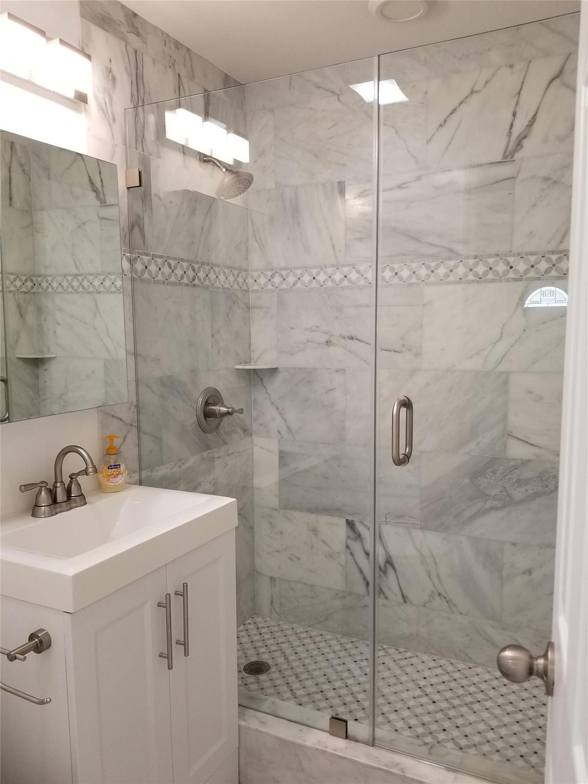 White shaker-style vanity with an integrated sink, brushed nickel faucet, frameless glass shower enclosure, shower walls featuring large-format tile and a decorative mosaic band, and a mosaic tile shower floor