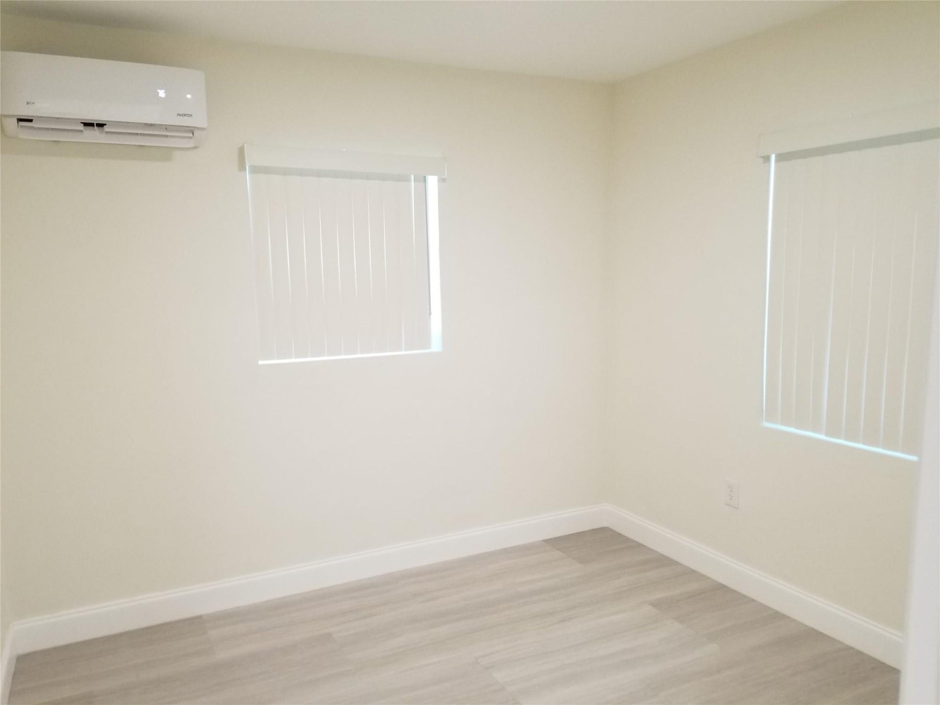 Window-mounted climate control unit, two windows with blinds, light-toned walls, and wood-finish flooring