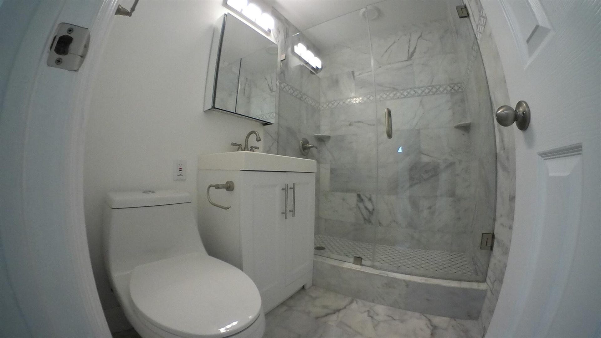 White vanity with a chrome faucet, frameless mirrored medicine cabinet, glass-enclosed shower, and full-height marble-style tile