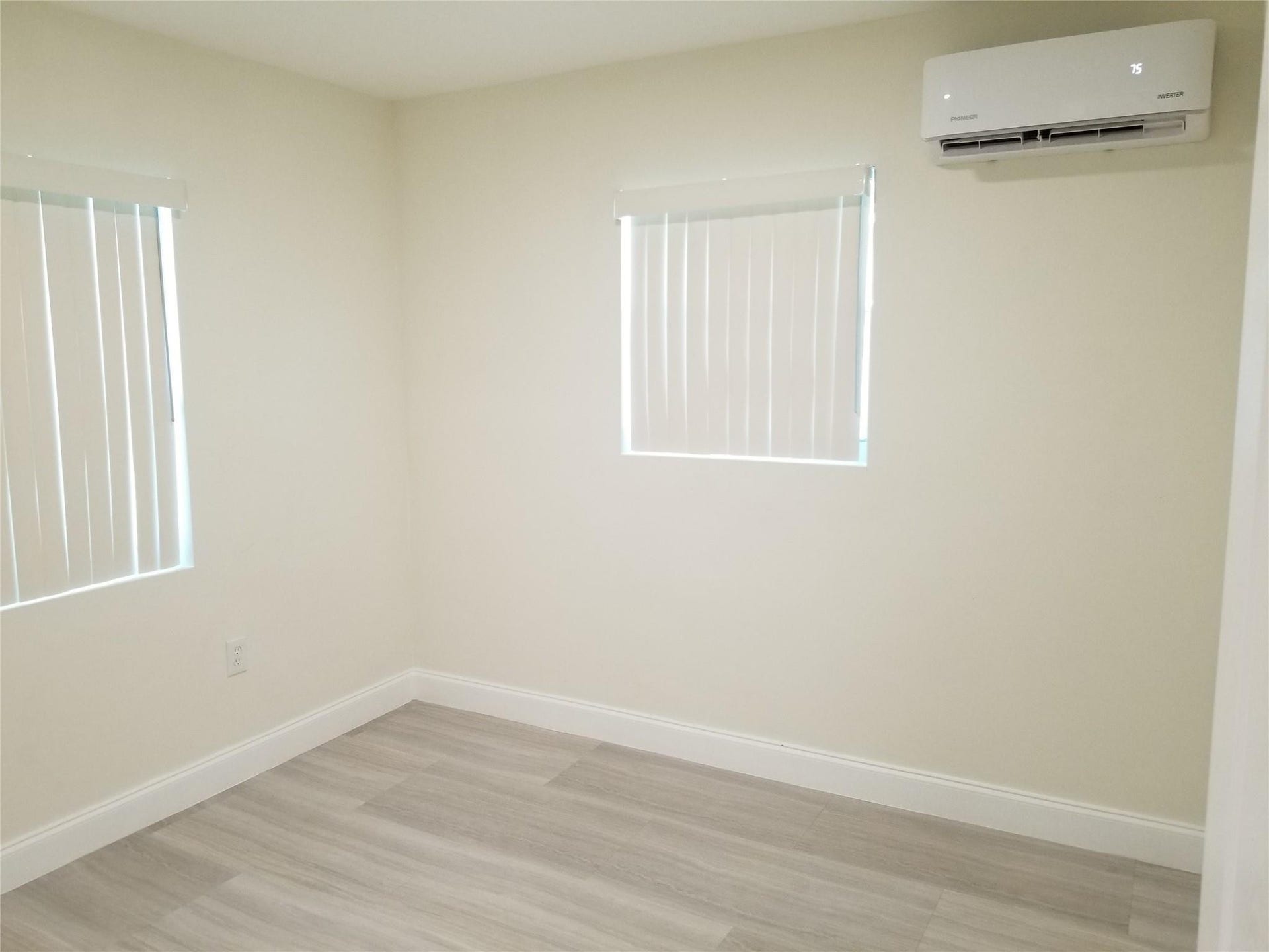 Light-toned wood-finish flooring, two windows with vertical blinds, and a wall-mounted air conditioning unit