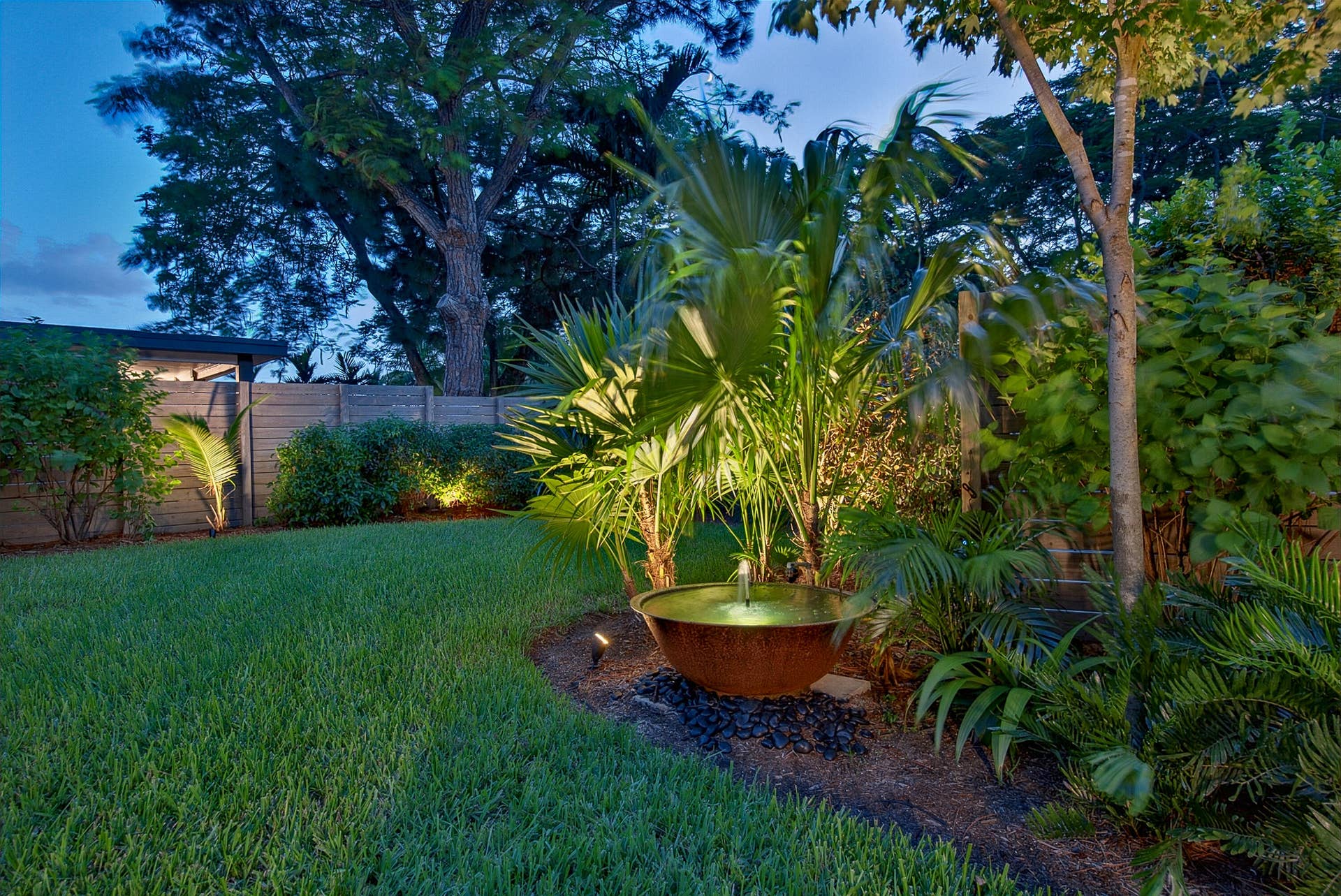 4850 NE 5TH Avenue, Oakland Park, FL 33334 The lighting highlights the manicured lawn, varied tropical landscaping, water feature, and mature trees