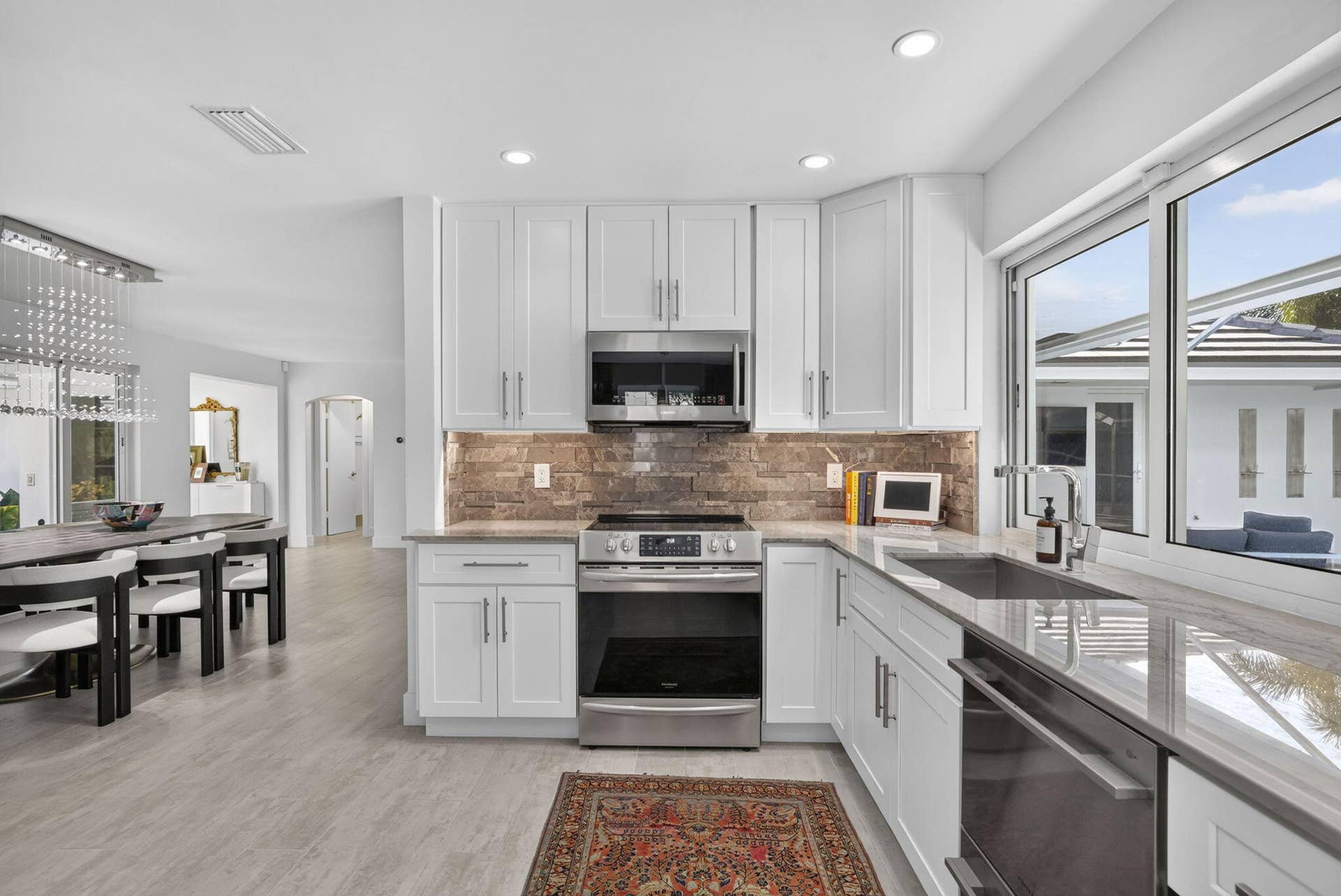 White shaker cabinetry, stainless steel appliances, stone tile backsplash, light-toned flooring, and large window