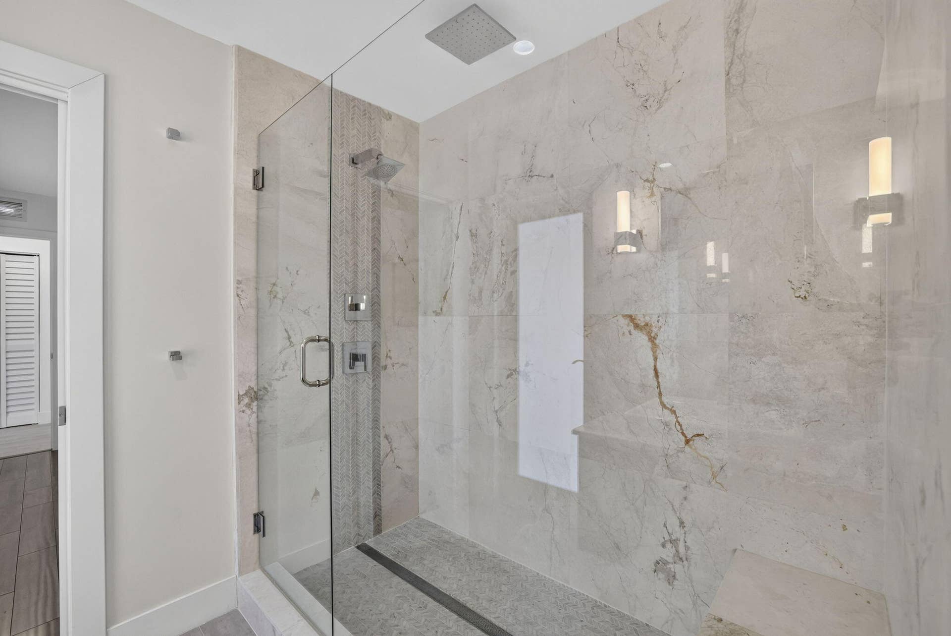 Frameless glass shower enclosure with a built-in bench, ceiling-mounted rain shower head, and wall-mounted sconce lighting