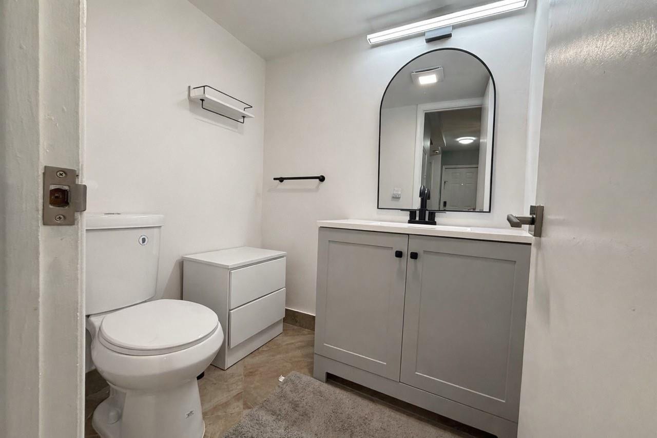 809 W Oakland Park Boulevard, #K4, Wilton Manors, FL 33311 White walls, tile flooring, grey vanity with double cabinet doors, arched mirror, and white toilet