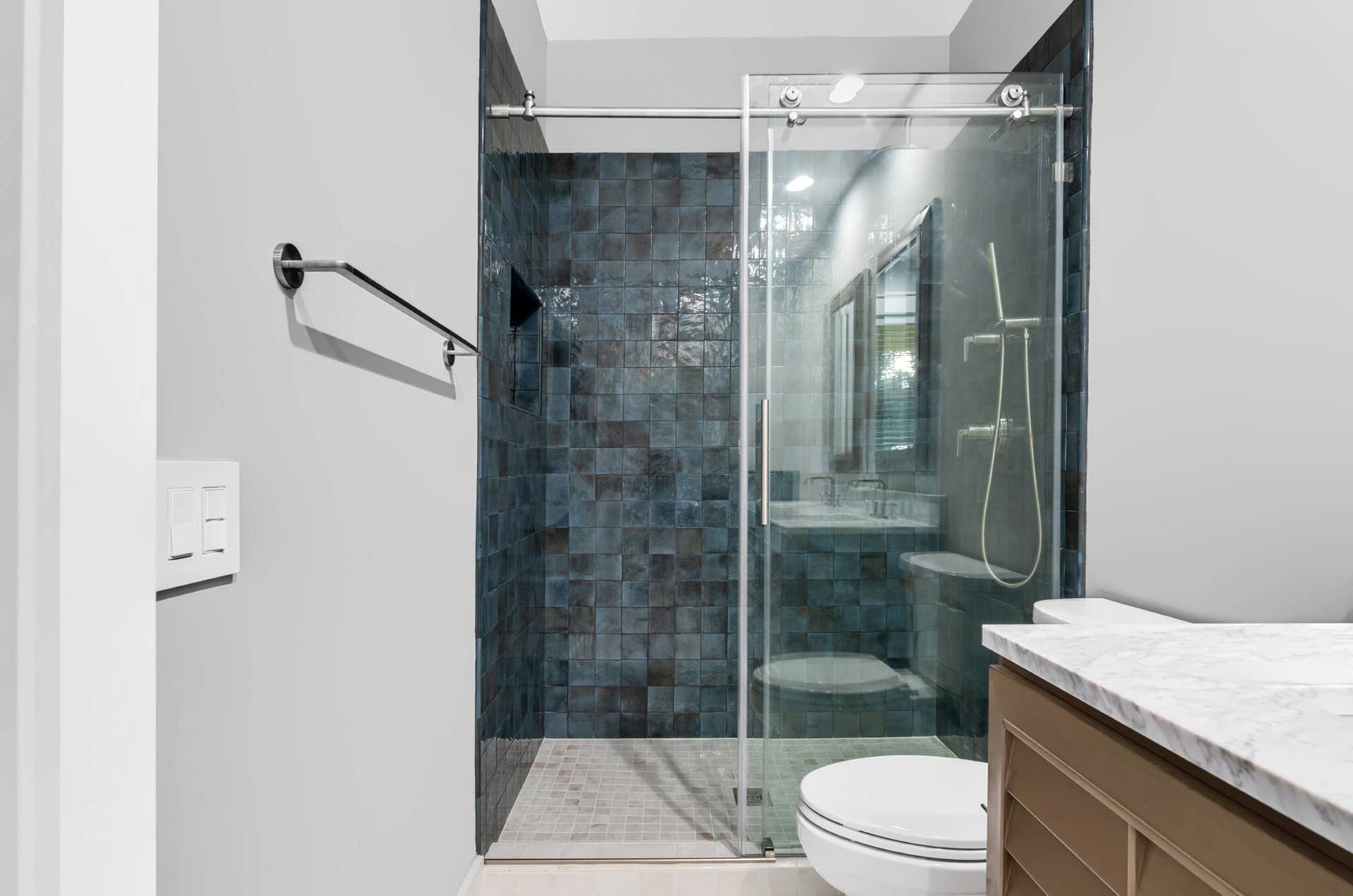 3207 E Community Drive E, Jupiter, FL 33458 Modern bathroom featuring a frameless glass shower enclosure with sliding glass doors, dark tile accent wall, white subway tile flooring, white ceramic toilet, and a wood-finish vanity with a white quartz countertop and integrated sink