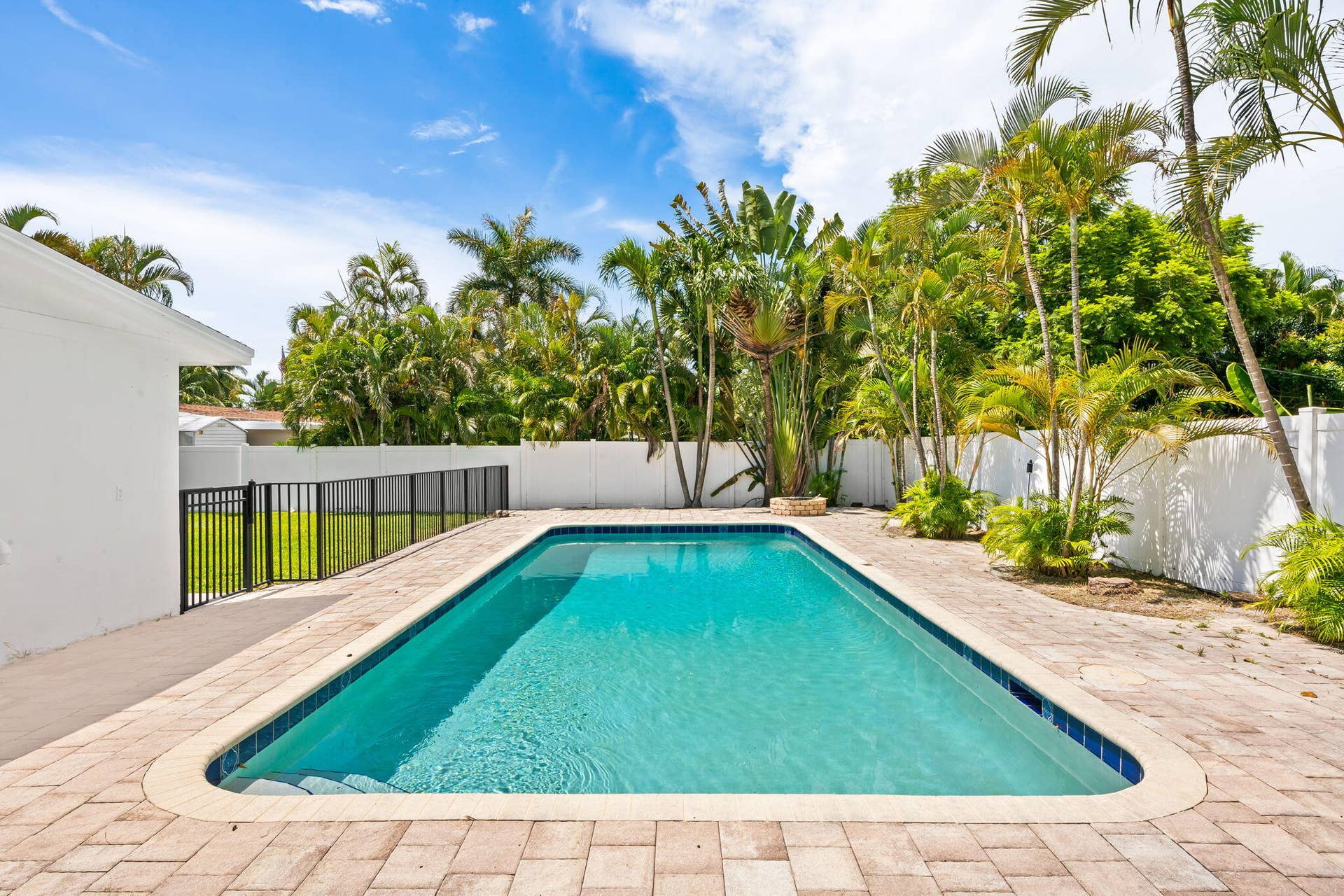 Rectangular swimming pool with curved corners, paver decking, and a black metal fence enclosing a grass area