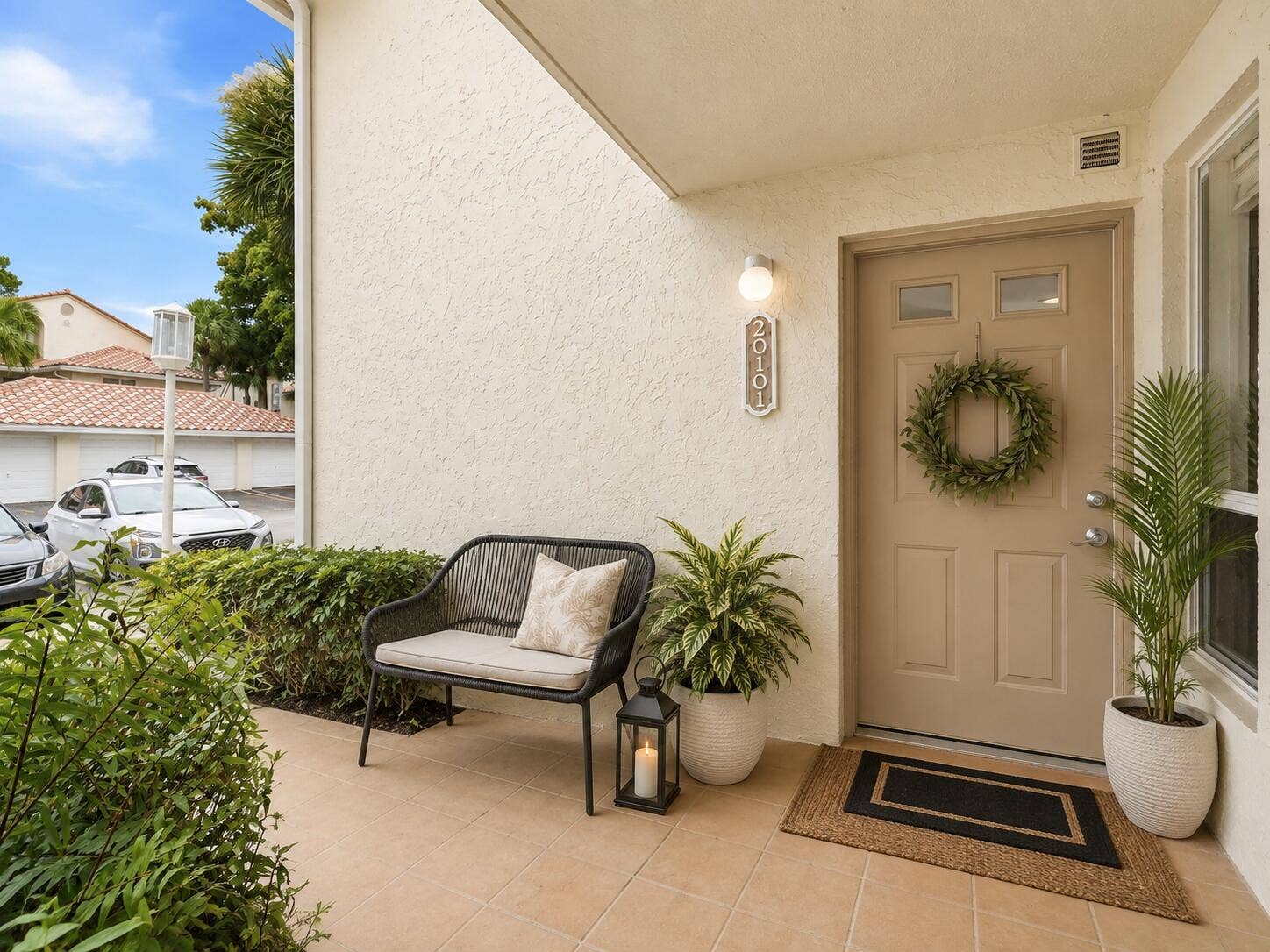 20101 Glenmoor Drive, West Palm Beach, West Palm Beach, FL 33409 Welcoming South Florida condo entrance with a front door, outdoor bench, potted tropical plants, lantern, wreath, and layered welcome mat.