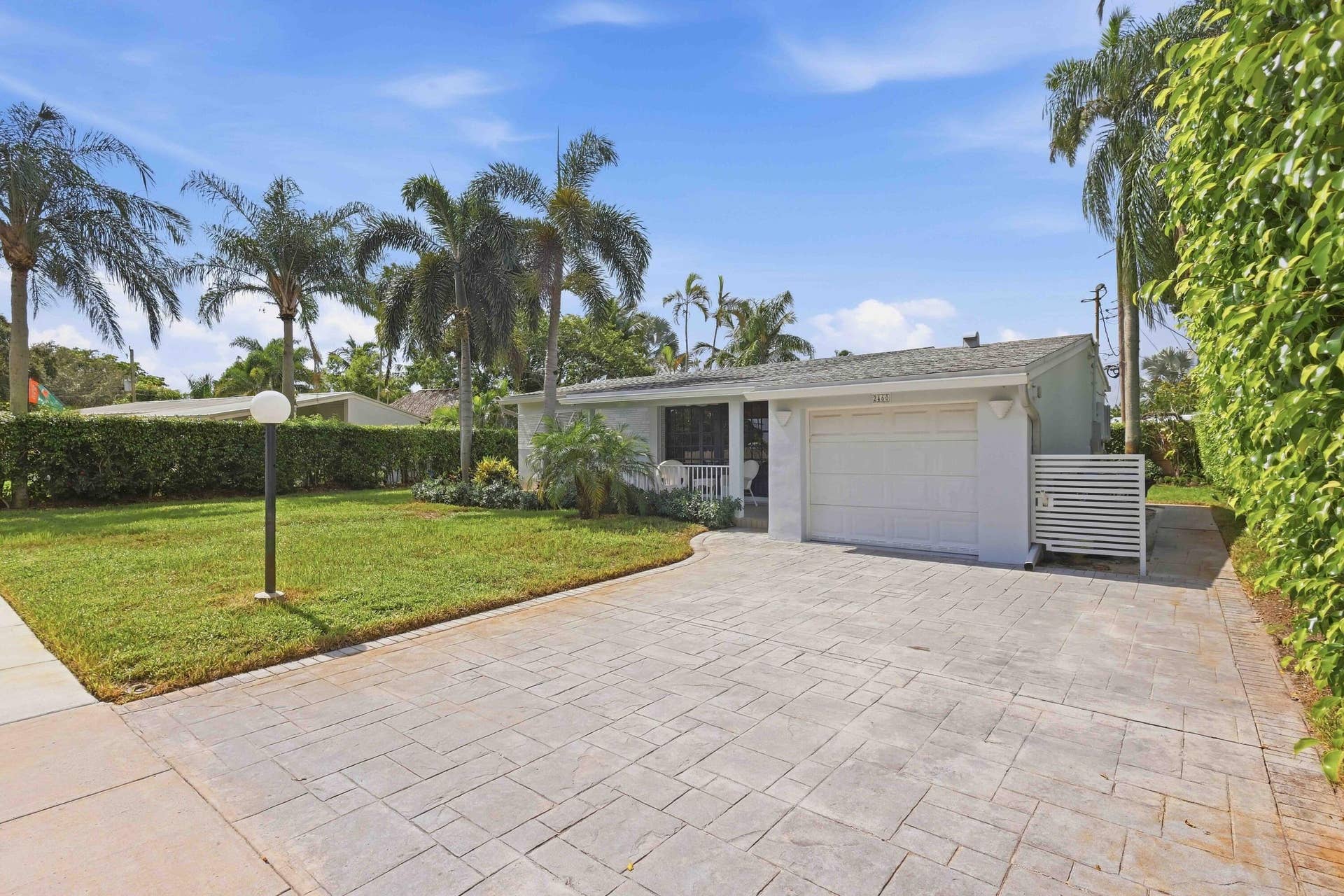 Stamped concrete driveway, attached garage, manicured lawn, established palm trees, and privacy hedging
