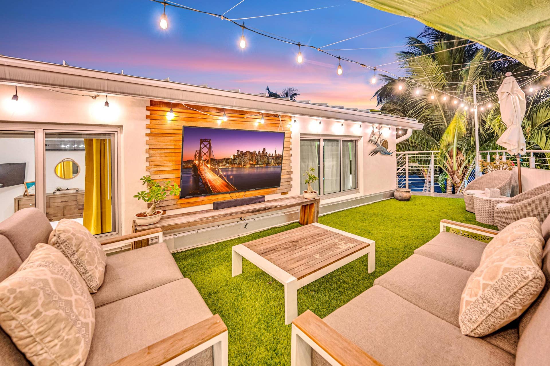 510 SW 18th Avenue, Fort Lauderdale, FL 33312 Outdoor entertainment space featuring a manicured lawn, string lighting overhead, built-in seating with wood-finish frames and cushioned upholstery, a wood-finish coffee table, and an integrated entertainment wall with a large flat-screen display and warm accent lighting