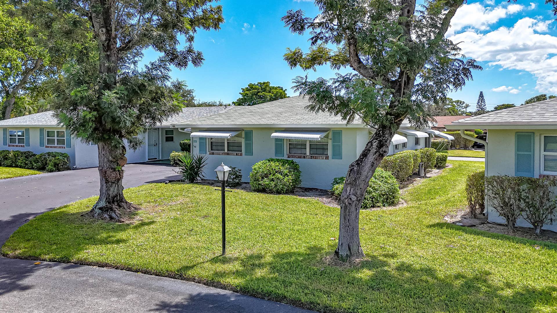 450 BLUEBIRD Lane, Delray Beach, FL 33445 Light-toned exterior with blue shutters, white awnings, mature trees, manicured lawns, and asphalt driveways