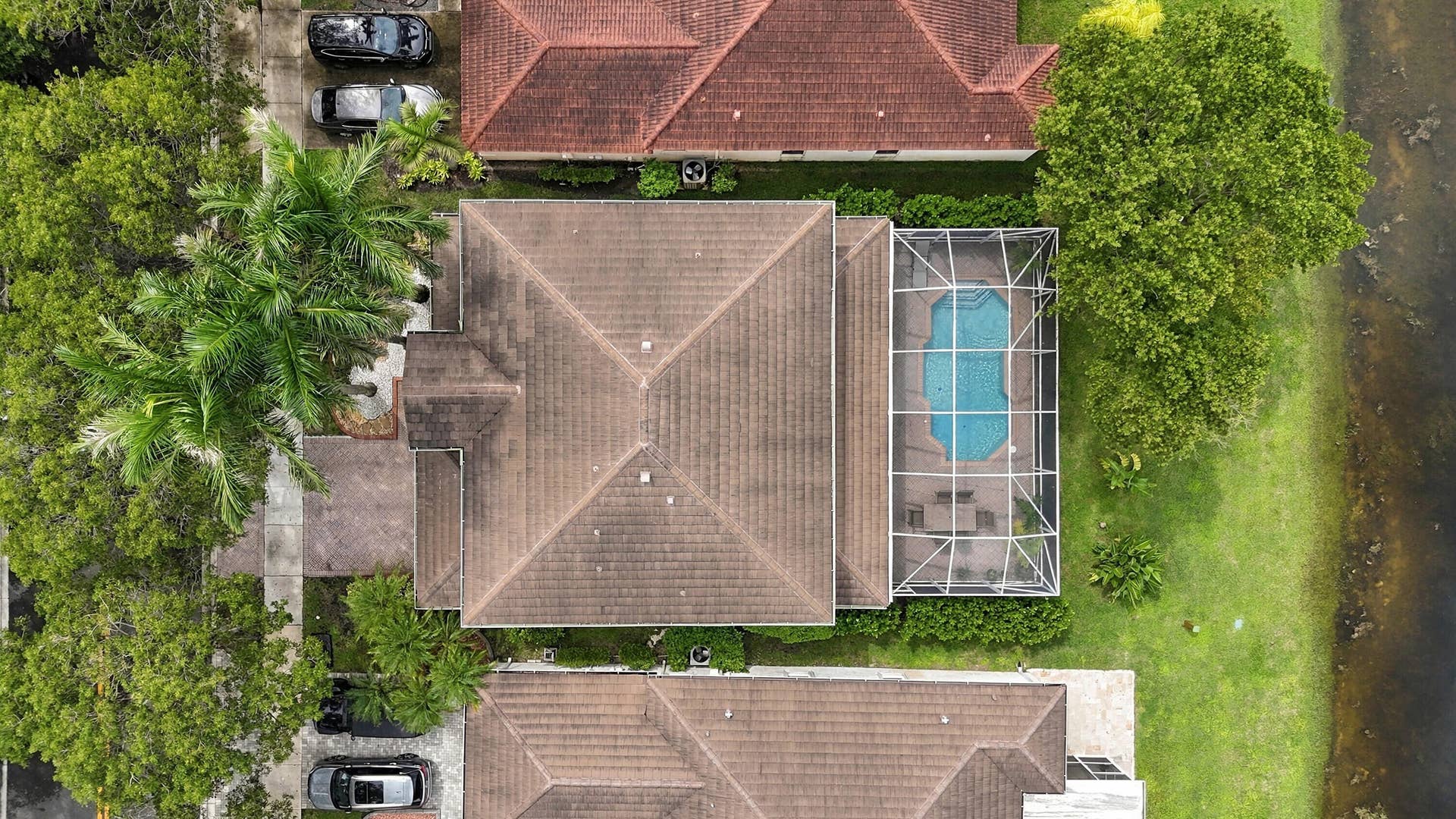1020 Lavender Circle, Weston, FL 33327 Terracotta tile roof with multiple gables, screened-in swimming pool, patio area, mature landscaping, and waterfront property with natural bank
