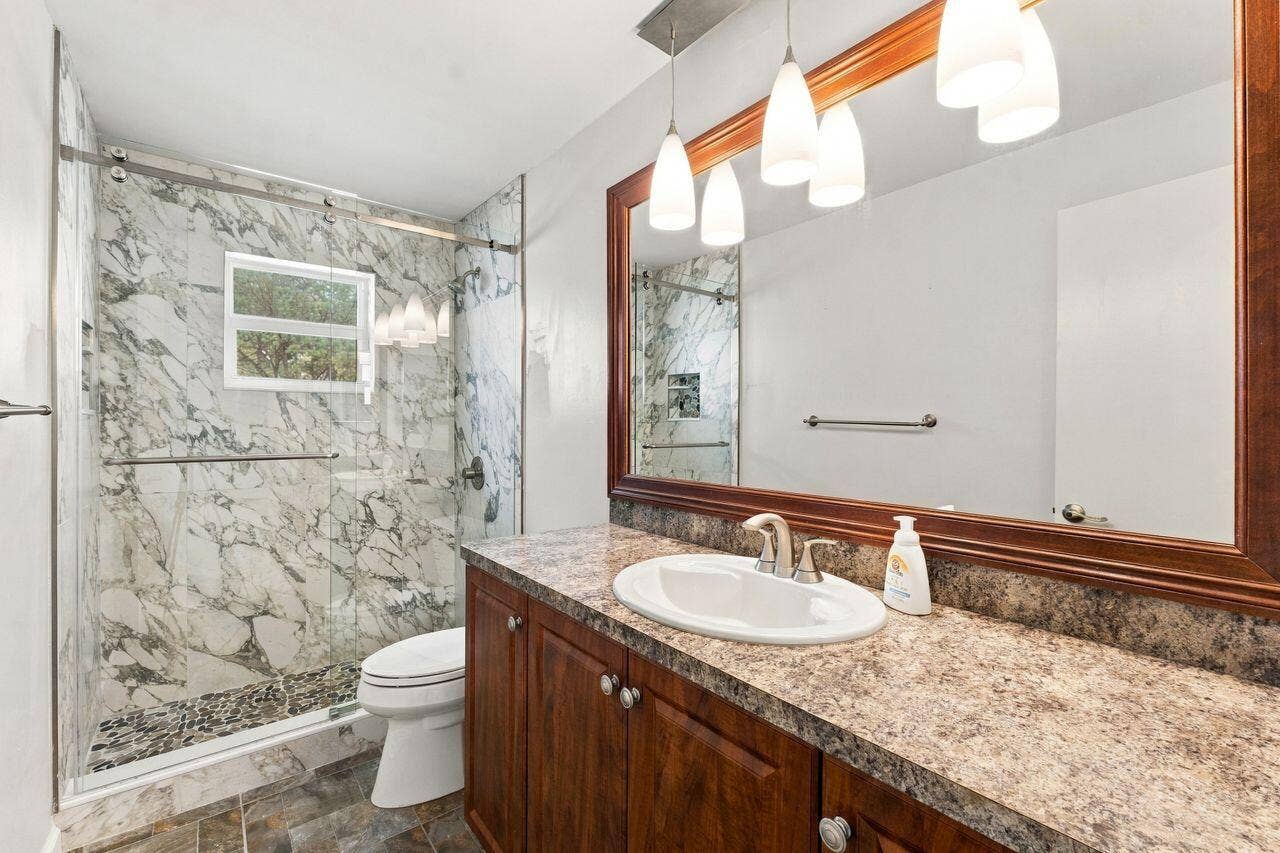 1003 SW 17th Street, Boynton Beach, FL 33426 Walk-in shower with marble-style tile surround and pebbled floor, vanity with wood-finish cabinetry, stone-look countertop, and large framed mirror