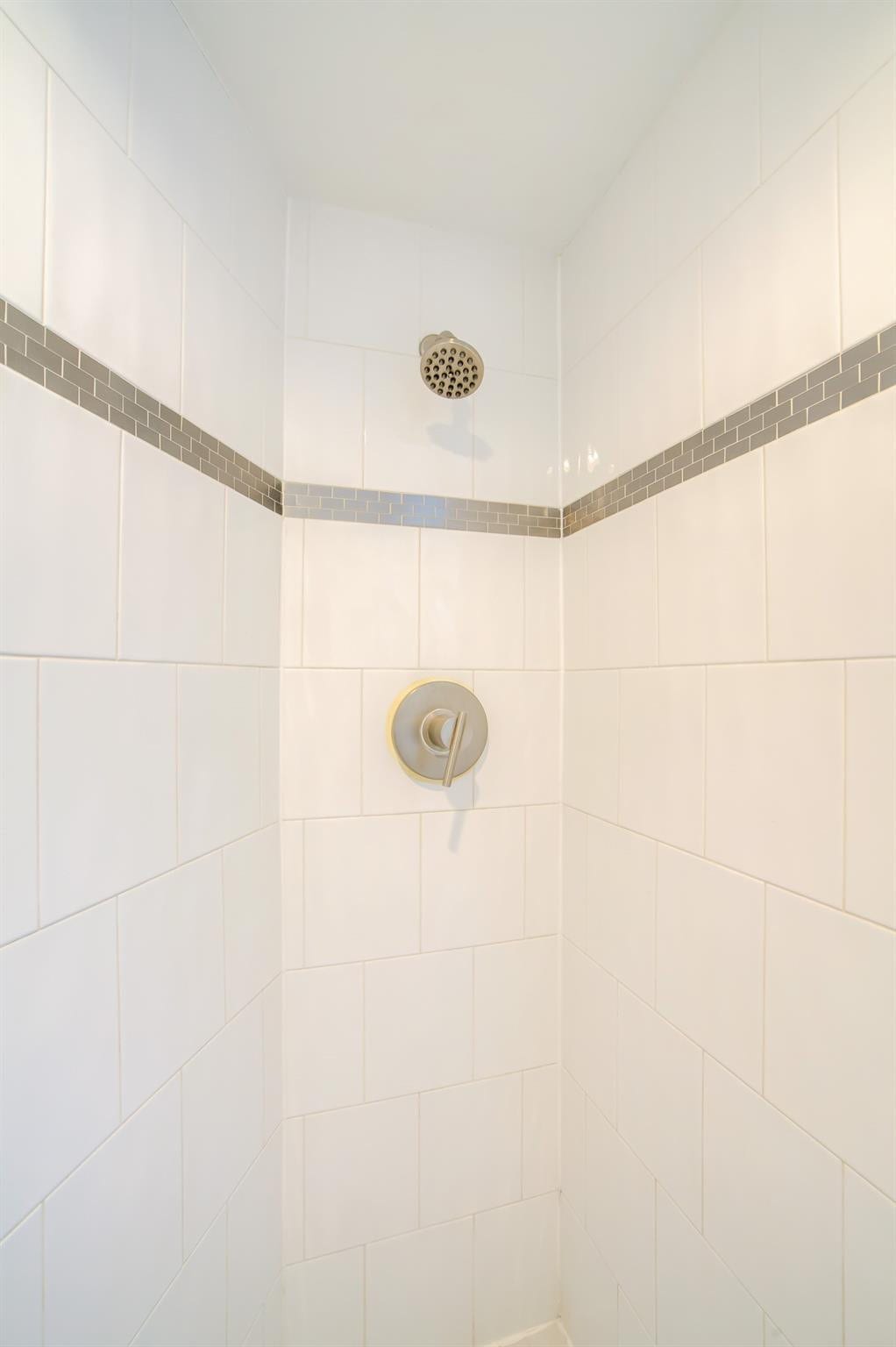 Shower enclosure with white tile walls, a decorative gray mosaic tile accent band, and a brushed nickel shower head and control