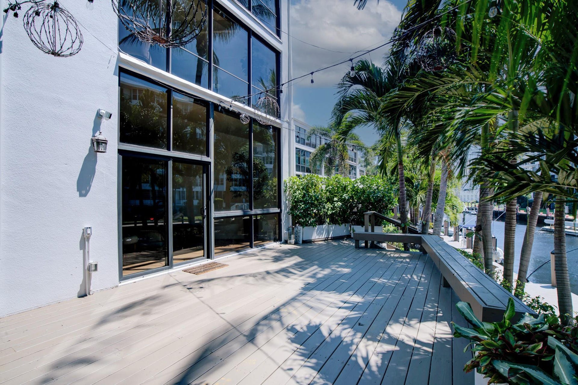 Waterfront property featuring a spacious outdoor deck with integrated seating, lush tropical landscaping, and expansive glass facade