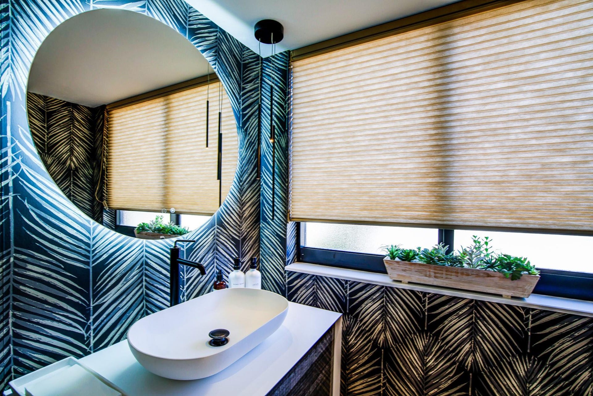 Round backlit mirror, oval vessel sink, black wall-mounted faucet, vanity with white countertop, and patterned wall covering