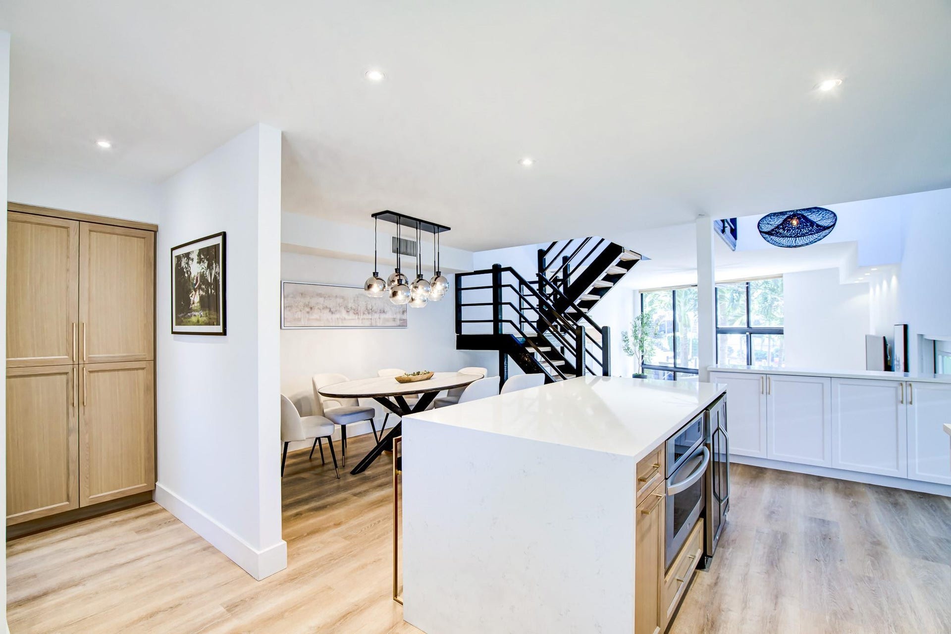 Open-concept living area featuring wood-finish flooring, a kitchen island with a built-in microwave, white cabinetry, and a contemporary black metal staircase