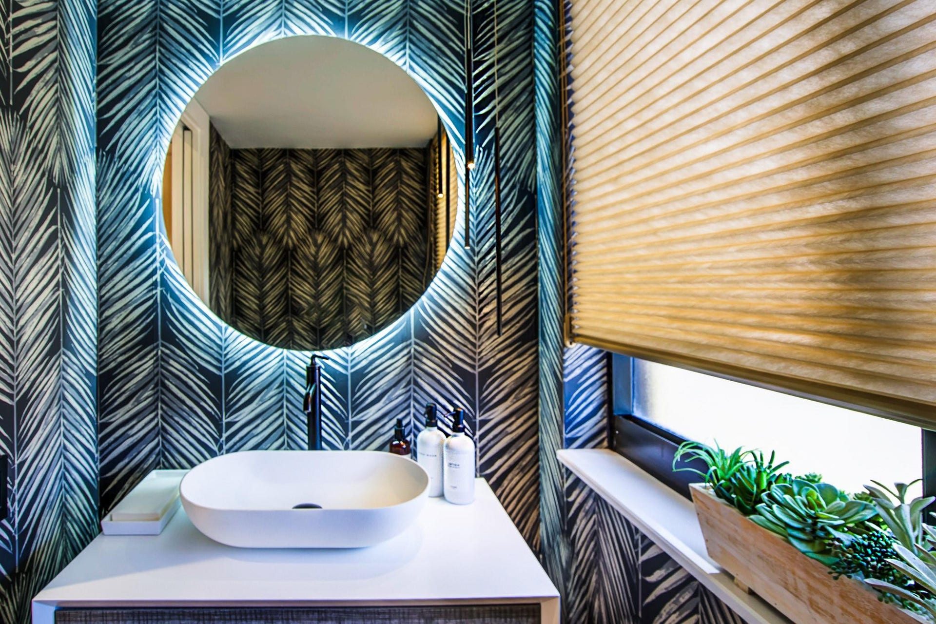 Backlit circular mirror, vessel sink, wall-mounted faucet, patterned wall covering, window with privacy blind