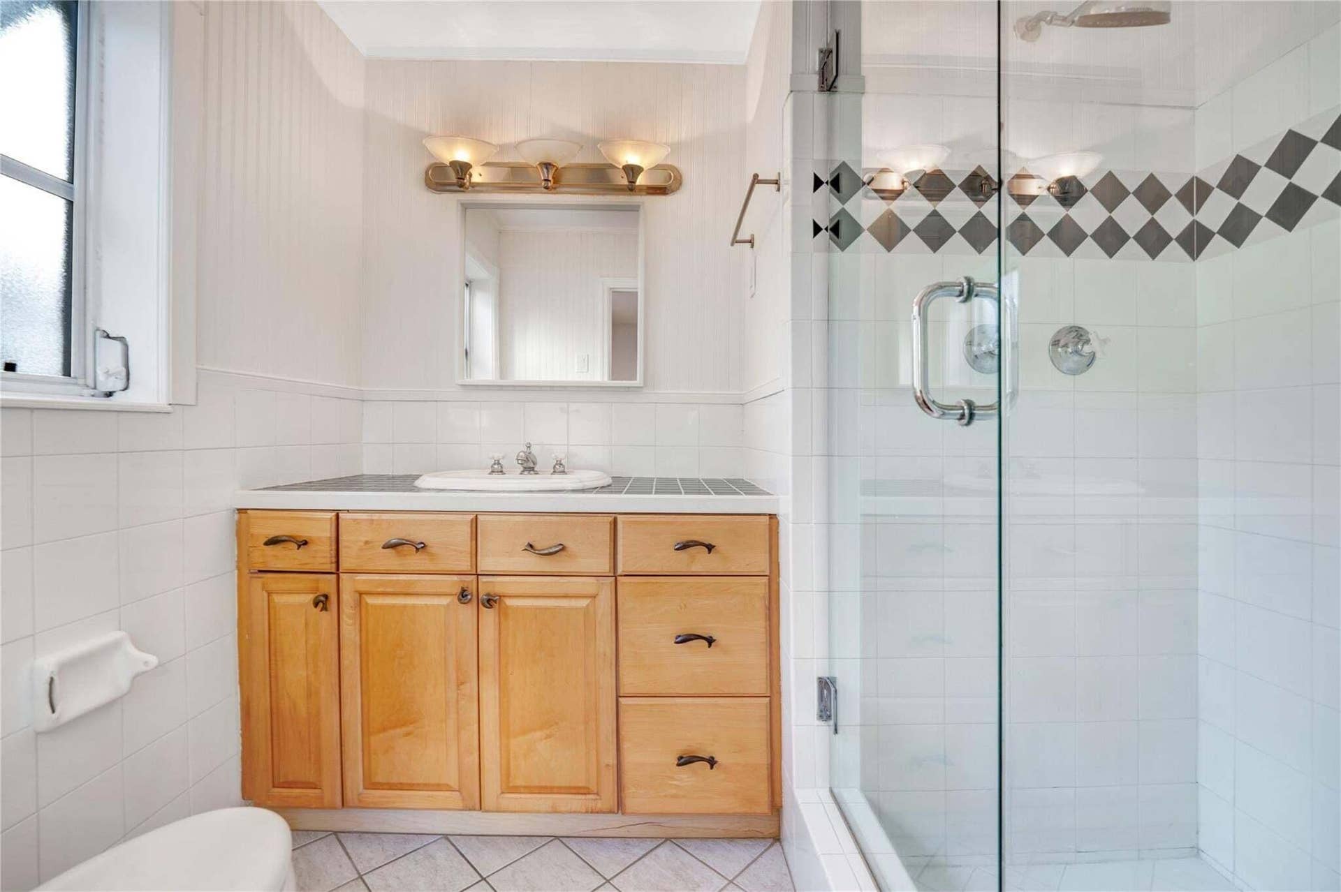 2142 NE 17th Avenue, Wilton Manors, FL 33305 White tiled bathroom featuring a wood-finish vanity with a built-in sink, a glass-enclosed shower with a decorative tile band, and a vanity light fixture