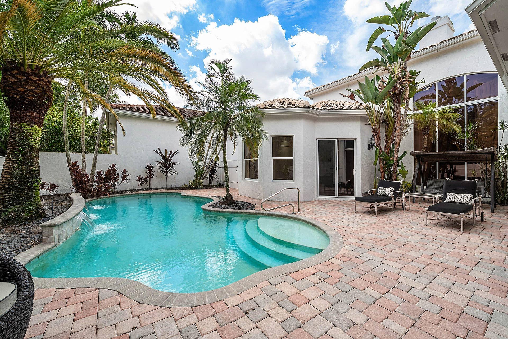 Curved swimming pool with waterfall feature, surrounded by a multi-tone paver patio and tropical landscaping