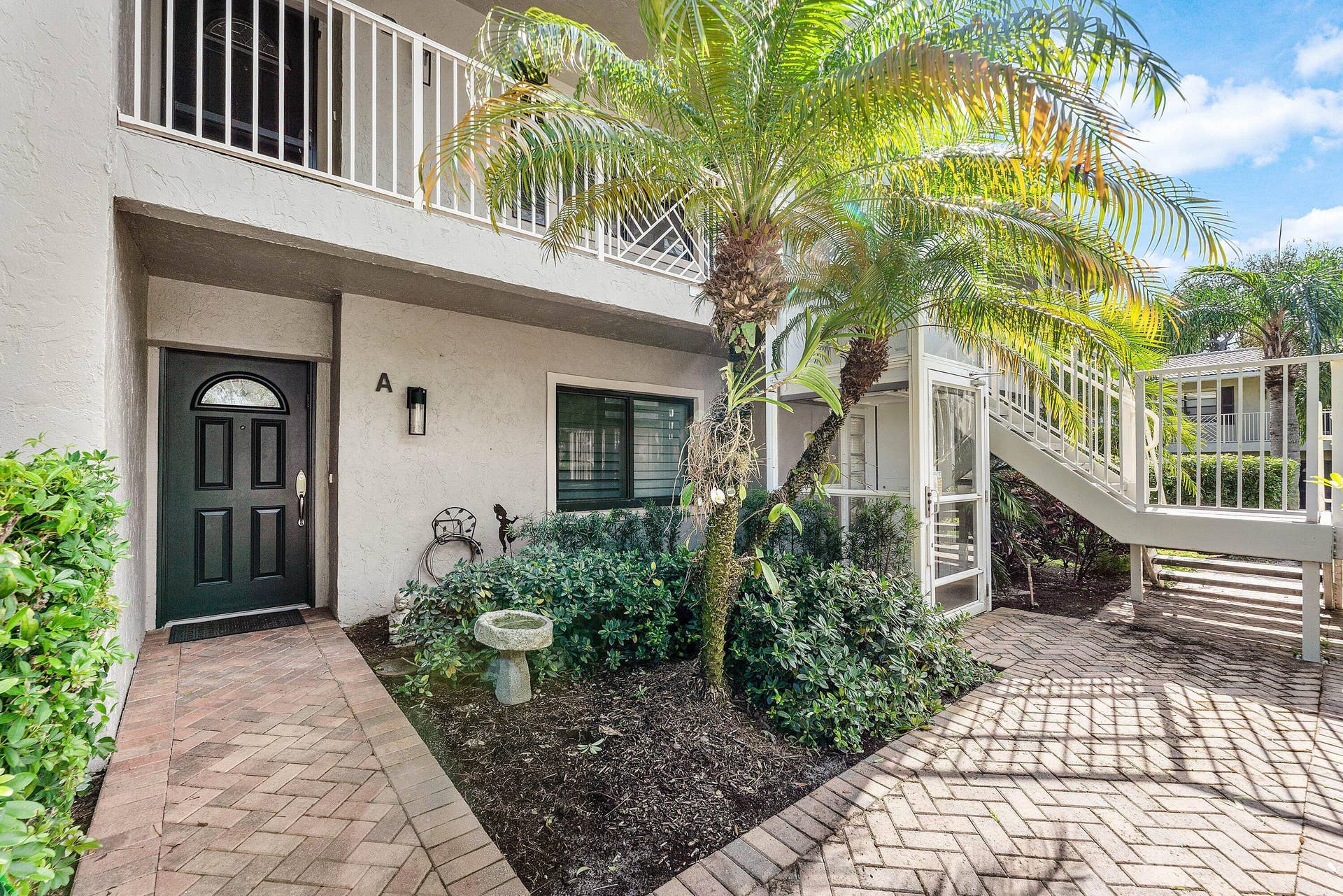 12 Stratford Drive E, #A, Boynton Beach, FL 33436 Brick paver walkway, dark paneled entry door with arched glass, exterior wall sconce, and mature landscaping