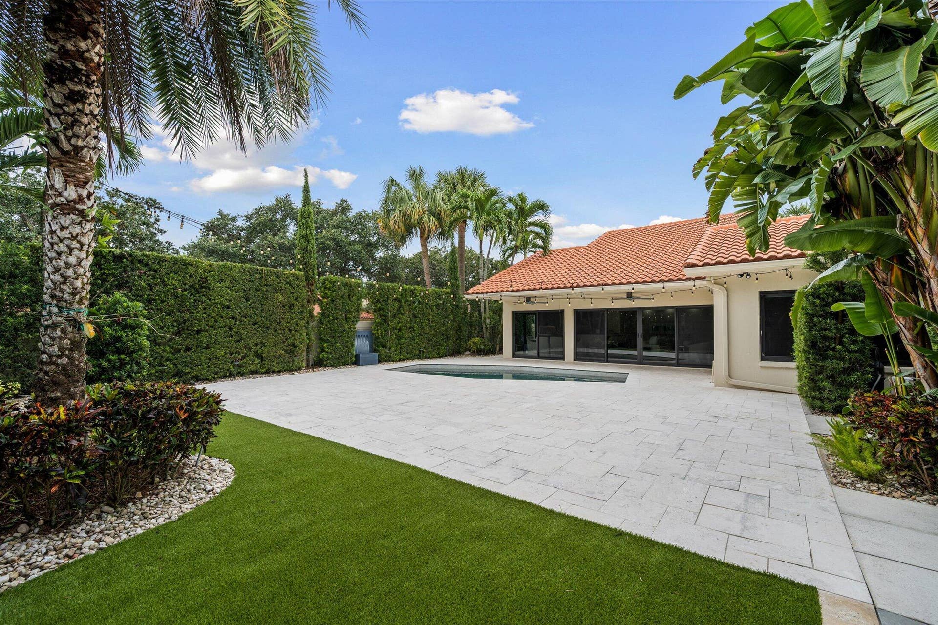 2890 ANTIETAM Lane, West Palm Beach, FL 33409 Expansive paver patio with a rectangular pool, artificial turf lawn, and mature tropical landscaping