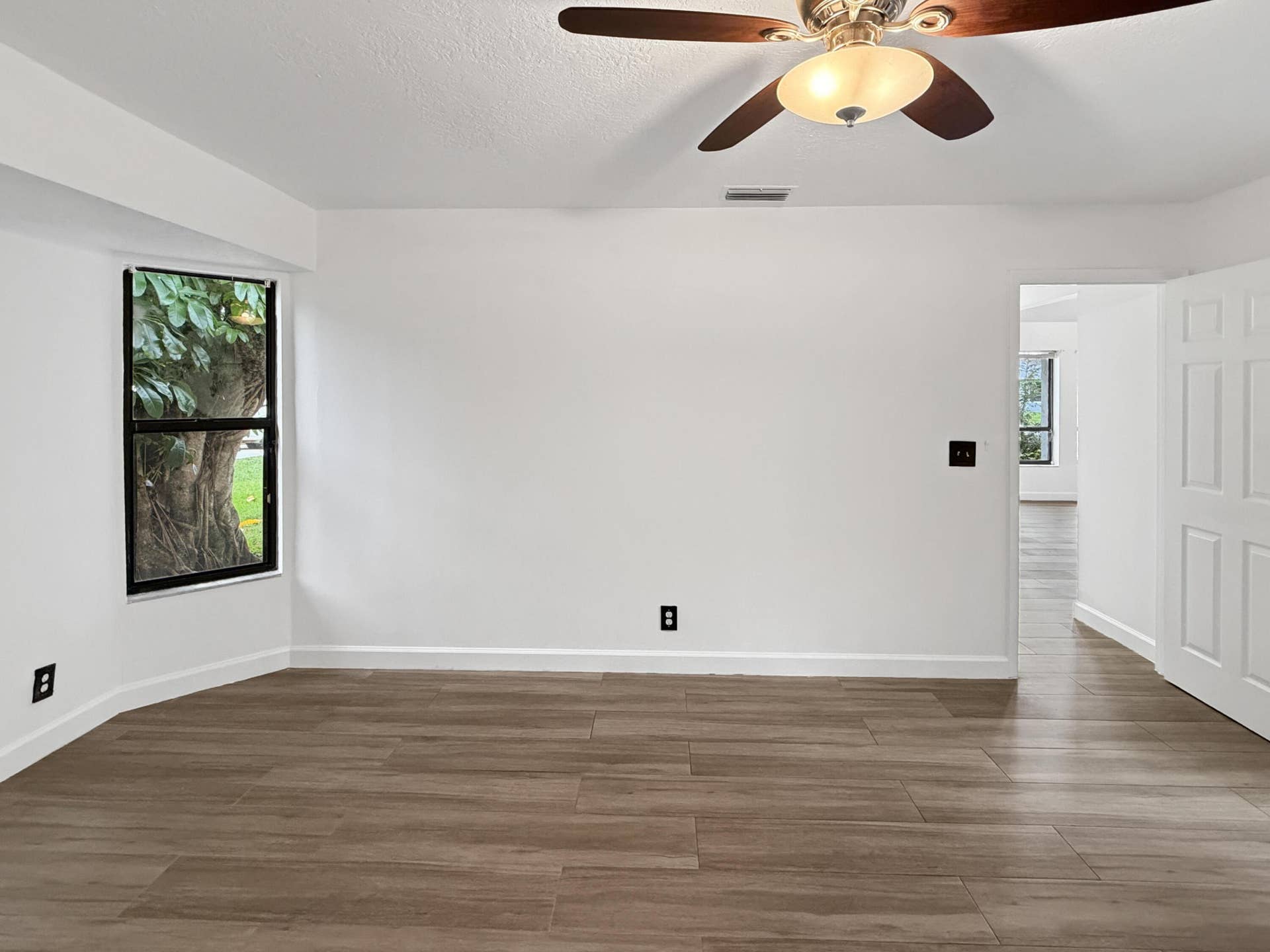 Wood-finish tile flooring, white painted walls, ceiling fan with integrated lighting, and a single window with exterior scenery