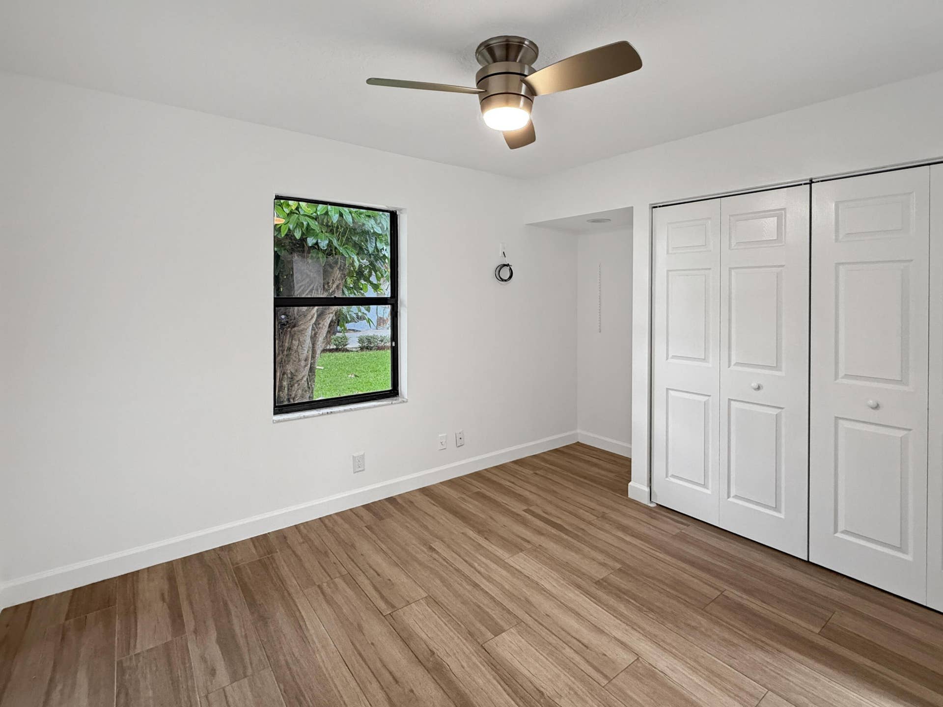 Spacious bedroom showcasing warm wood-finish flooring, white six-panel closet doors with multiple storage sections, ceiling-mounted fan with integrated lighting, and abundant natural light from a large window framing outdoor scenery