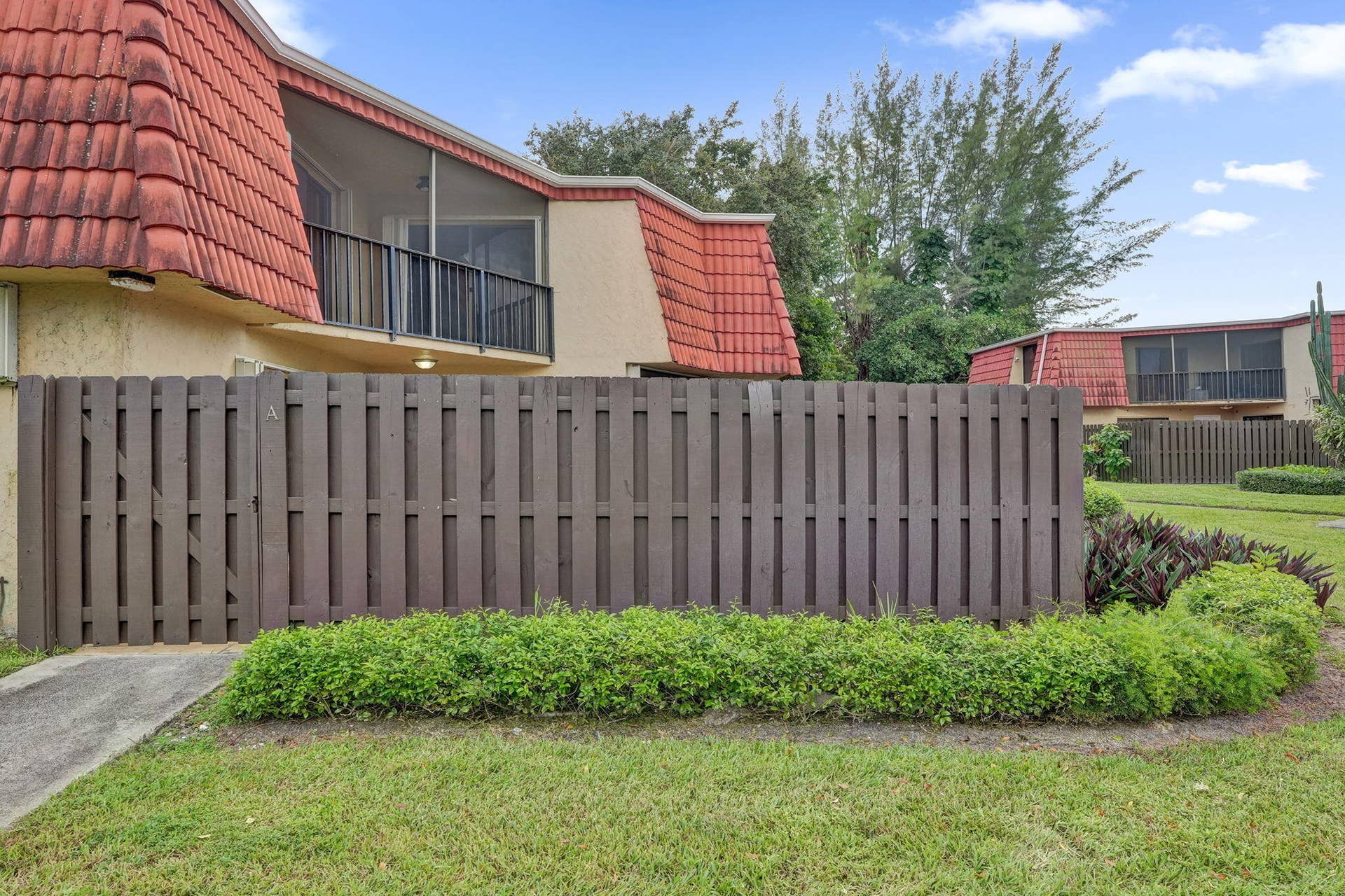 Terracotta-style roof tiles, stucco exterior, screened balcony with black railing, brown privacy fencing, and manicured landscaping
