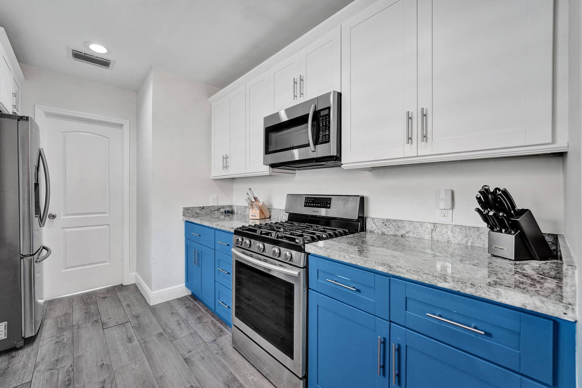 Modern kitchen featuring white cabinetry with stainless steel hardware, blue lower cabinet bases, gas range cooktop, built-in microwave, marble-look countertops, and wood-finish plank flooring throughout the space