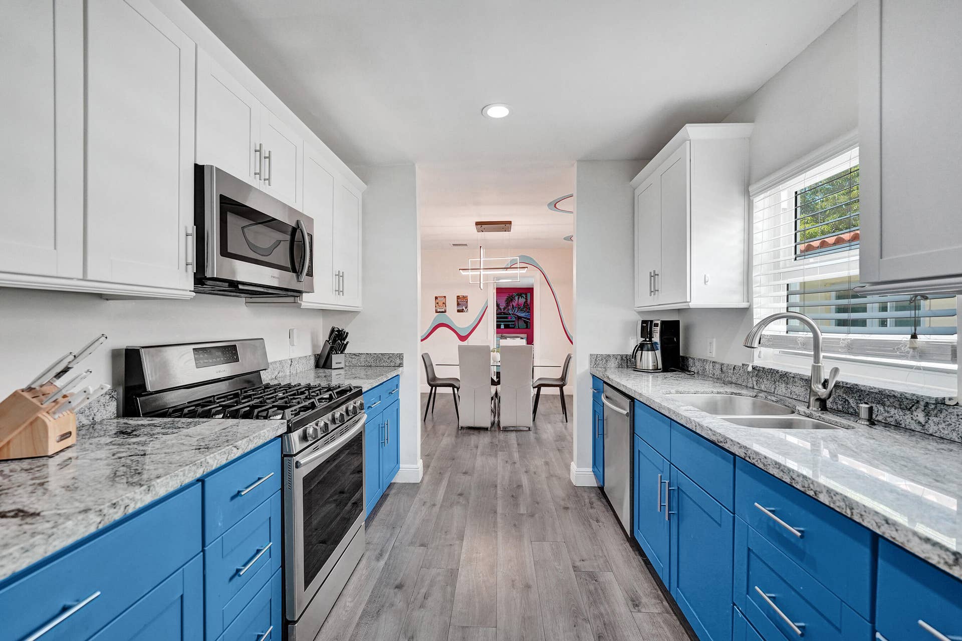 Galley kitchen featuring white upper cabinetry and blue lower cabinetry with brushed nickel hardware, granite countertops, stainless steel gas range with microwave above, undermount sink with chrome faucet, wood-finish flooring, and recessed lighting throughout