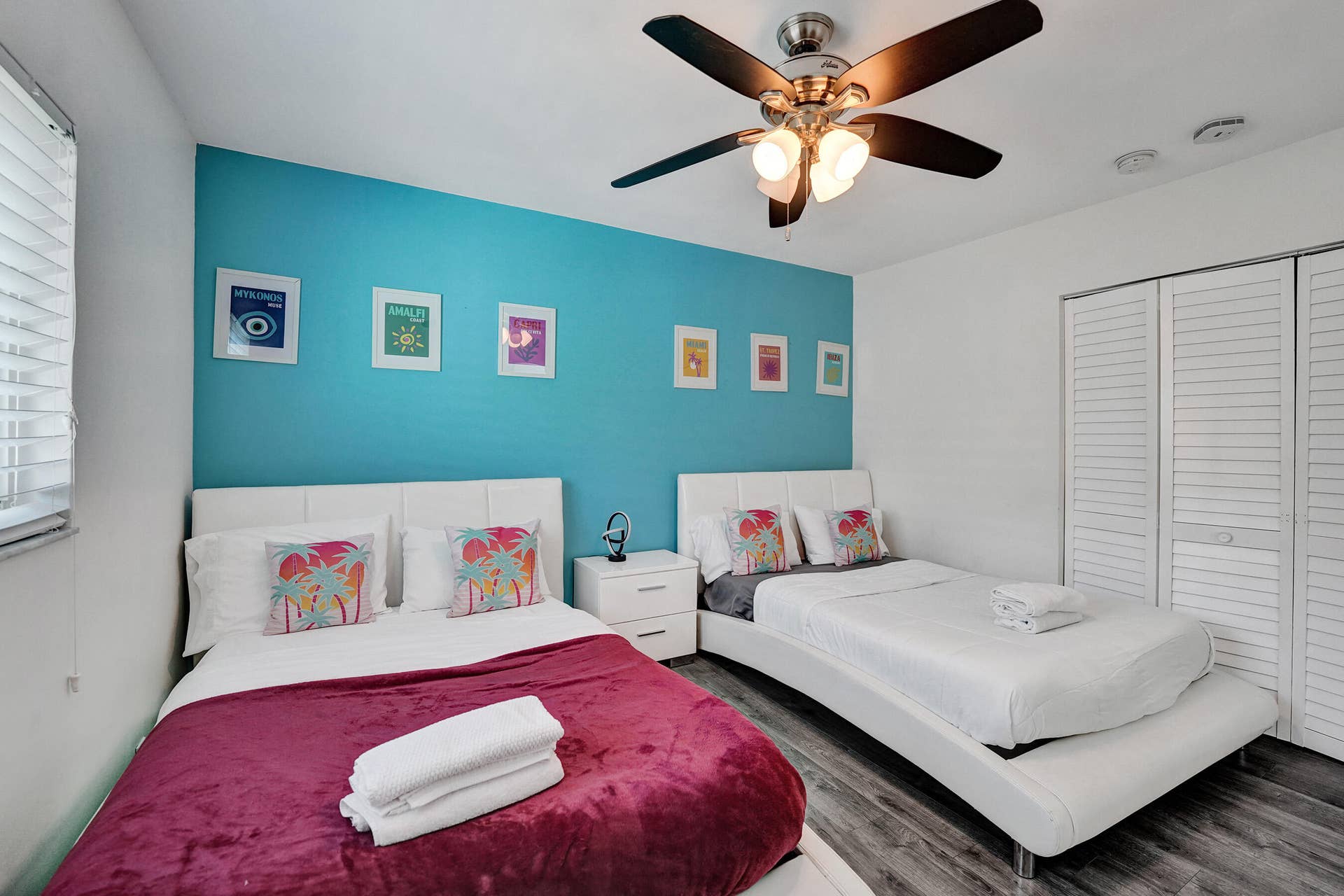 Wood-finish flooring with two white upholstered bed frames, a ceiling fan with integrated lighting, and white louvered bifold closet doors