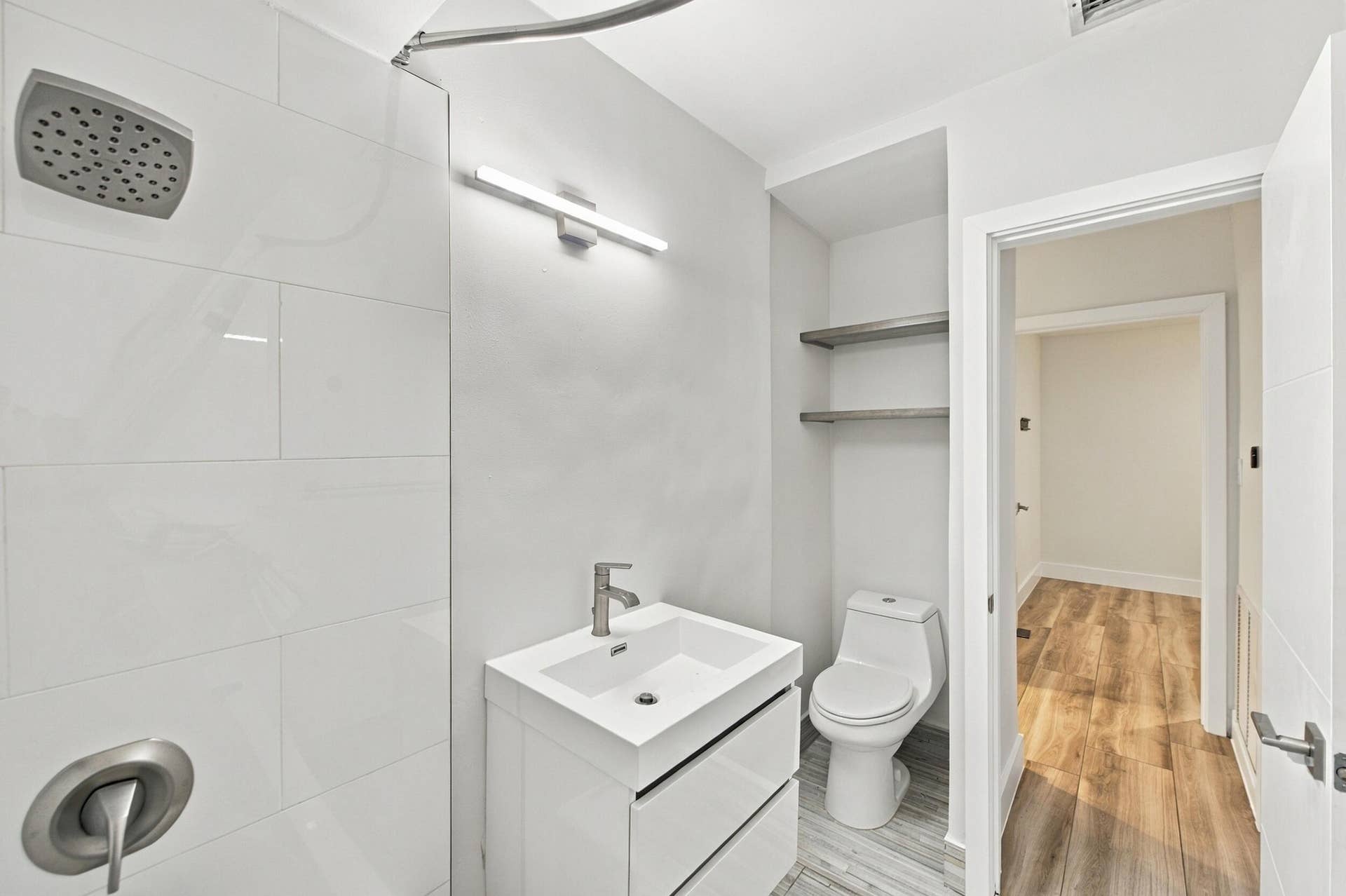 Walk-in shower with large format wall tiling, single vanity with integrated sink, modern wall-mounted light fixture, built-in shelving, and wood-finish flooring
