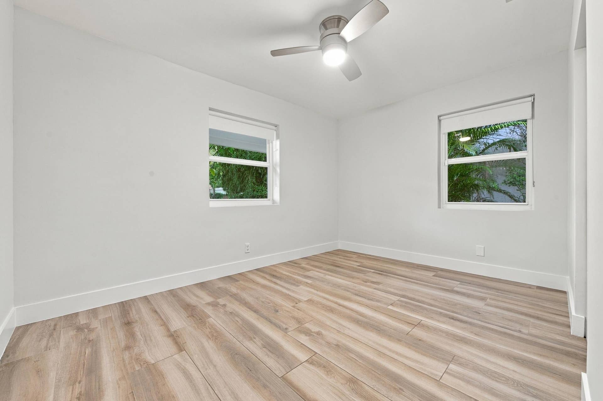 Light-toned wood-finish flooring, white walls, two windows with roller shades, and a ceiling fan with integrated lighting