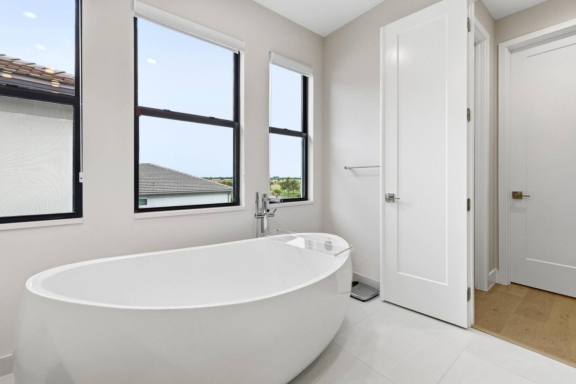 Freestanding soaking tub with floor-mounted faucet, complemented by large windows, white paneled doors, and light tile flooring