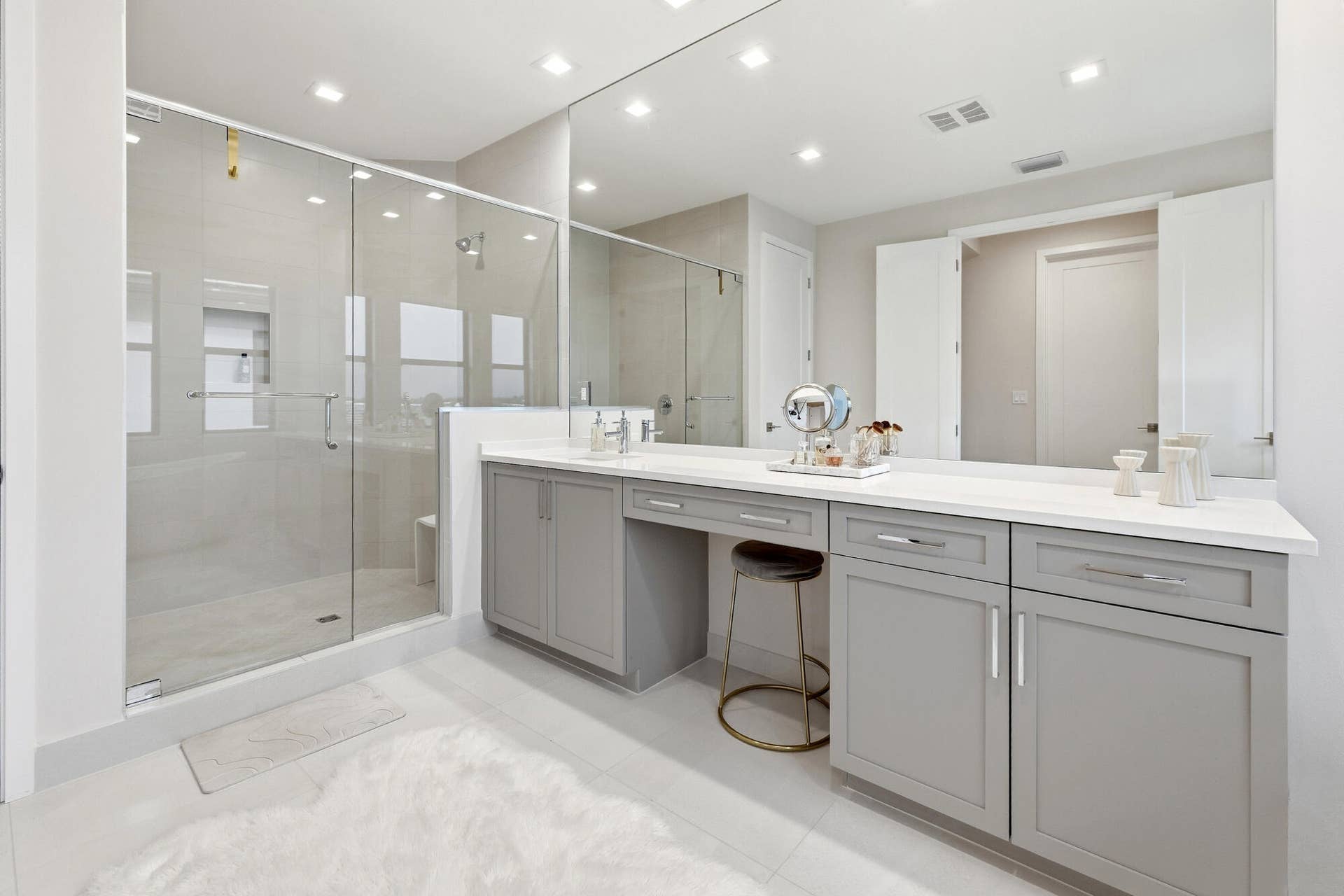 Spacious bathroom featuring a frameless glass shower enclosure, a full-width vanity mirror, light-toned tile flooring, recessed lighting, and gray shaker-style cabinetry with a built-in vanity area