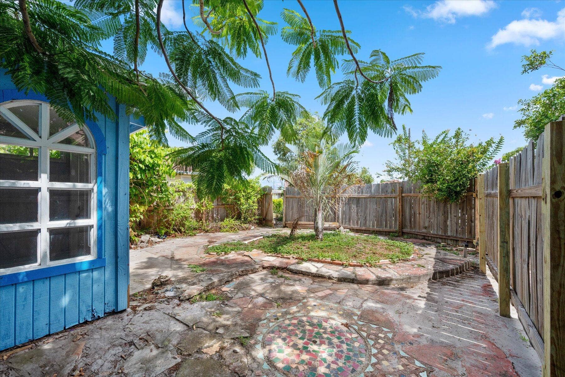 714 W Lakewood Road, #B, West Palm Beach, West Palm Beach, FL 33405 Enclosed backyard with a wooden fence, featuring a paved pathway, central planted area, and a blue exterior structure with an arched window