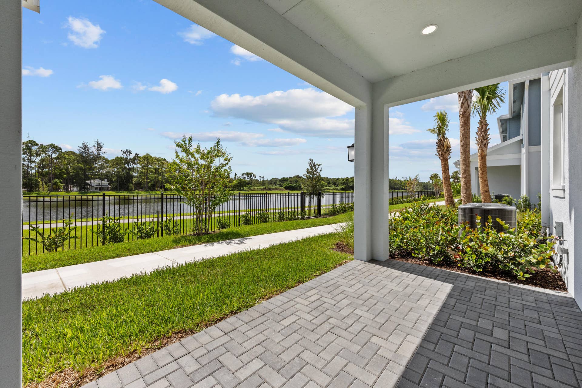 Covered patio with paver flooring, overlooking a water feature with a black metal fence, and featuring established landscaping