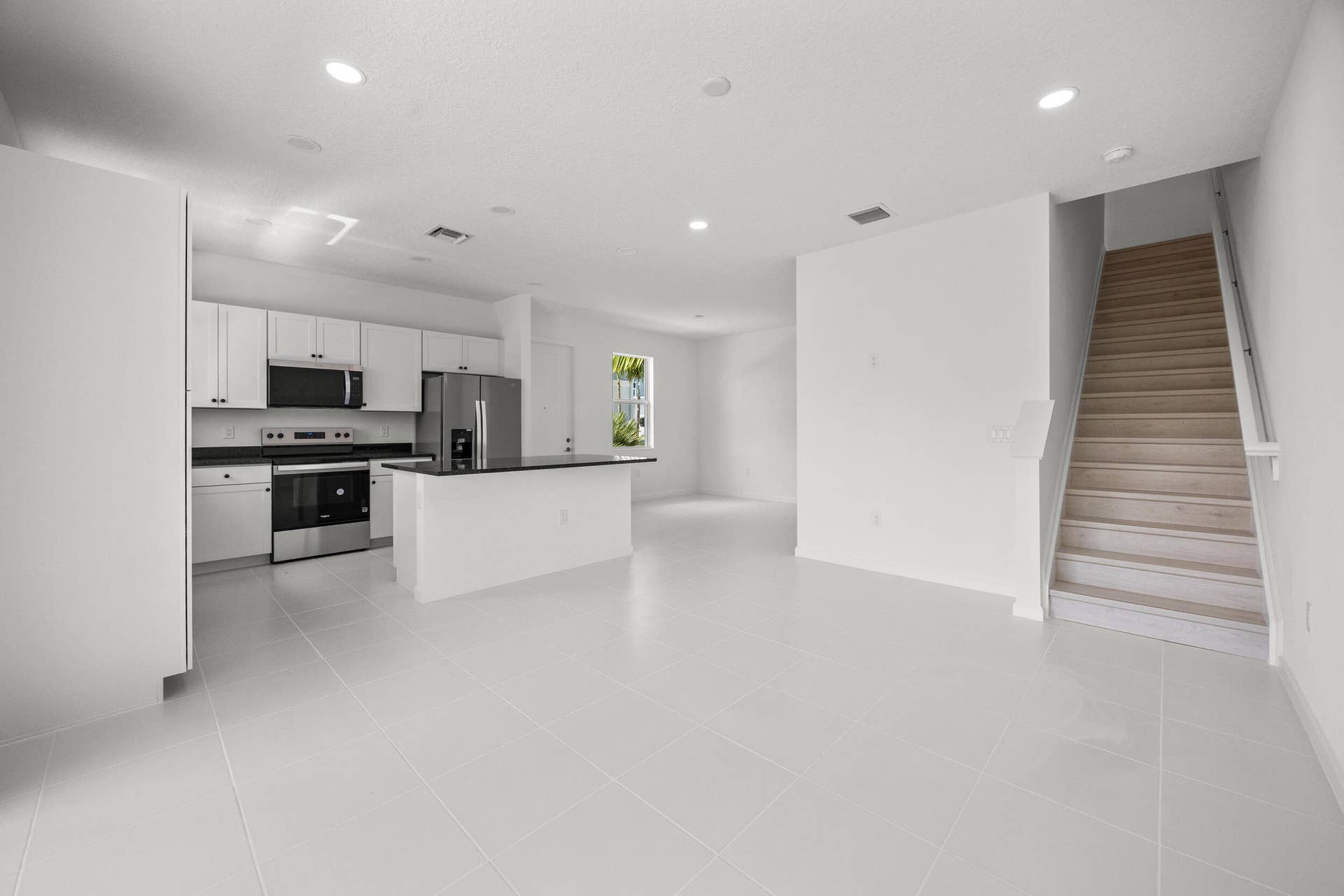 Open-concept living area with light-toned tiled flooring, white cabinetry, dark countertops, stainless steel appliances, and a wood staircase