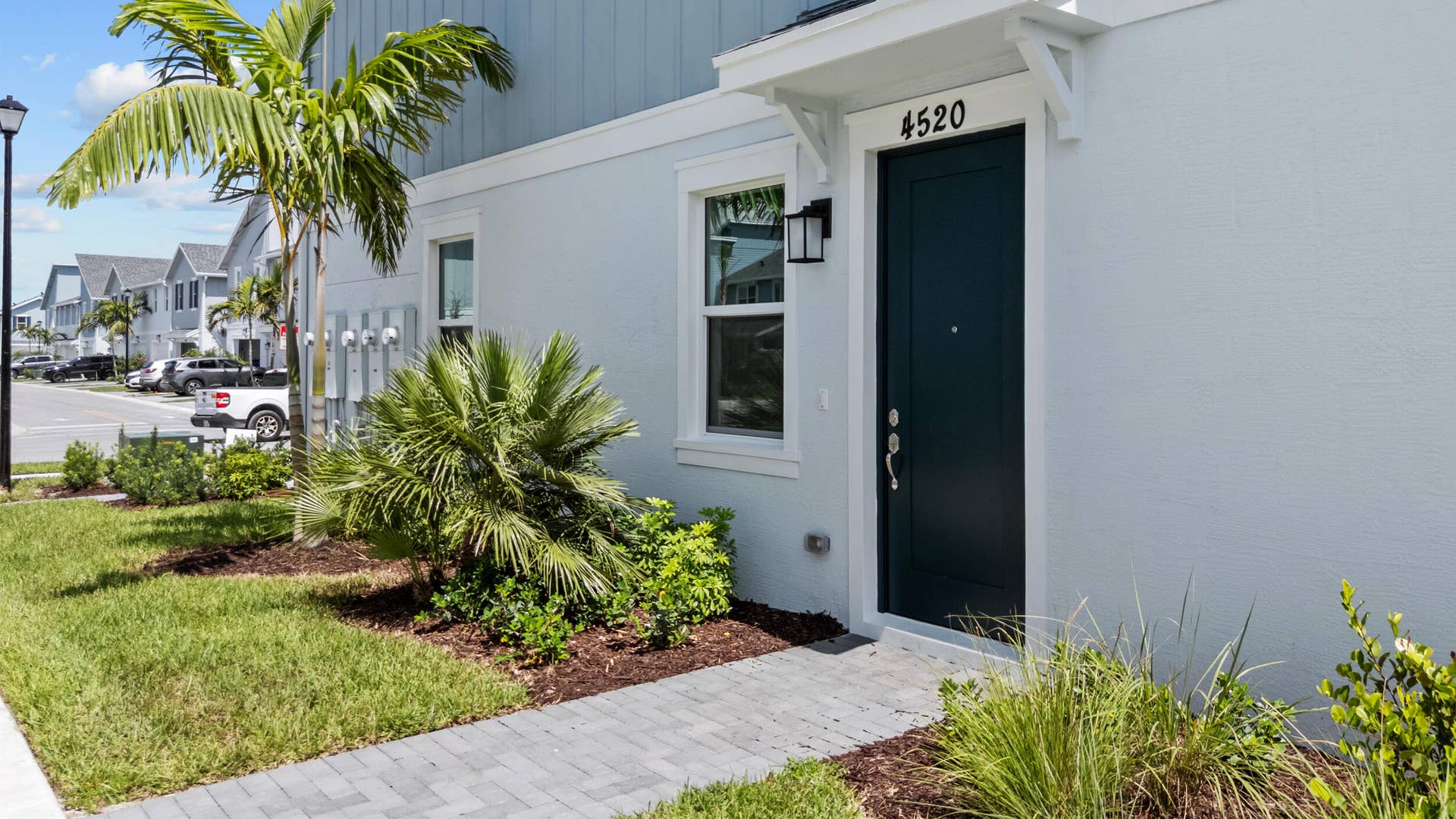 Paved walkway, vibrant green lawn, dark paneled entry door with white trim, exterior wall light fixture, and palm tree landscaping