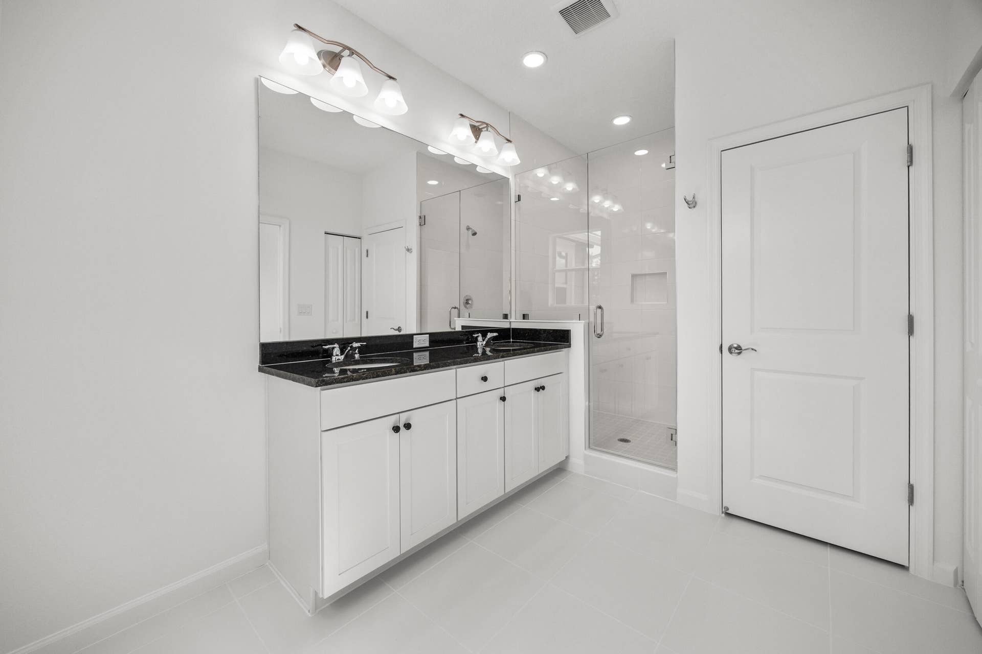 Dual vanity with dark countertop, large wall-mounted mirror, brushed nickel light fixtures, walk-in shower with glass enclosure, and white panel door