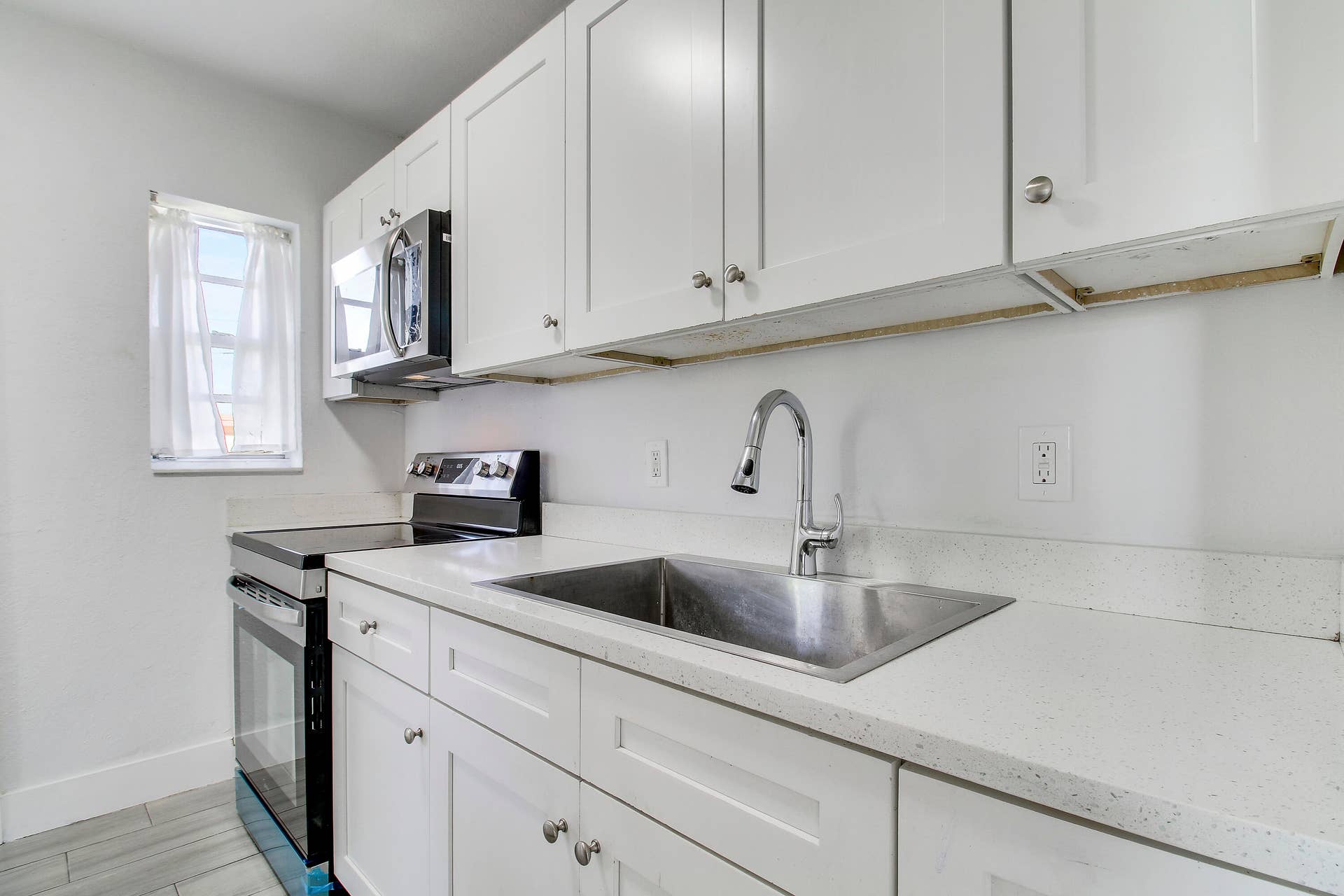 White shaker-style cabinetry with brushed nickel hardware, stainless steel undermount sink, contemporary gooseneck faucet, light-toned countertops, and tile flooring
